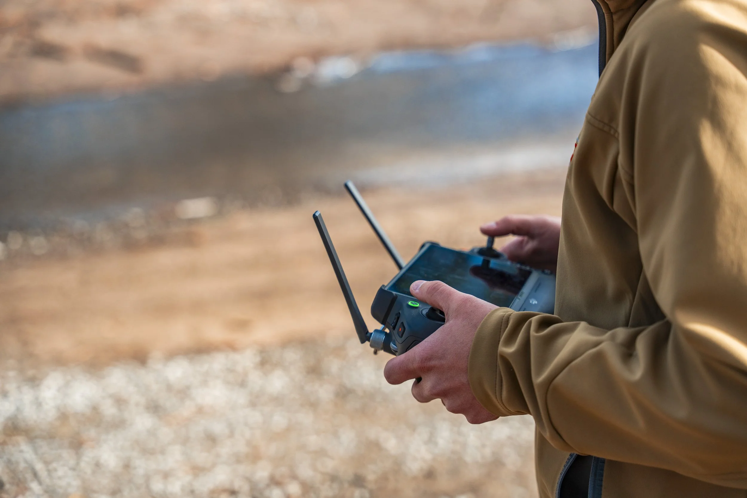 A person in a tan jacket operating a drone controller outdoors near a body of water.