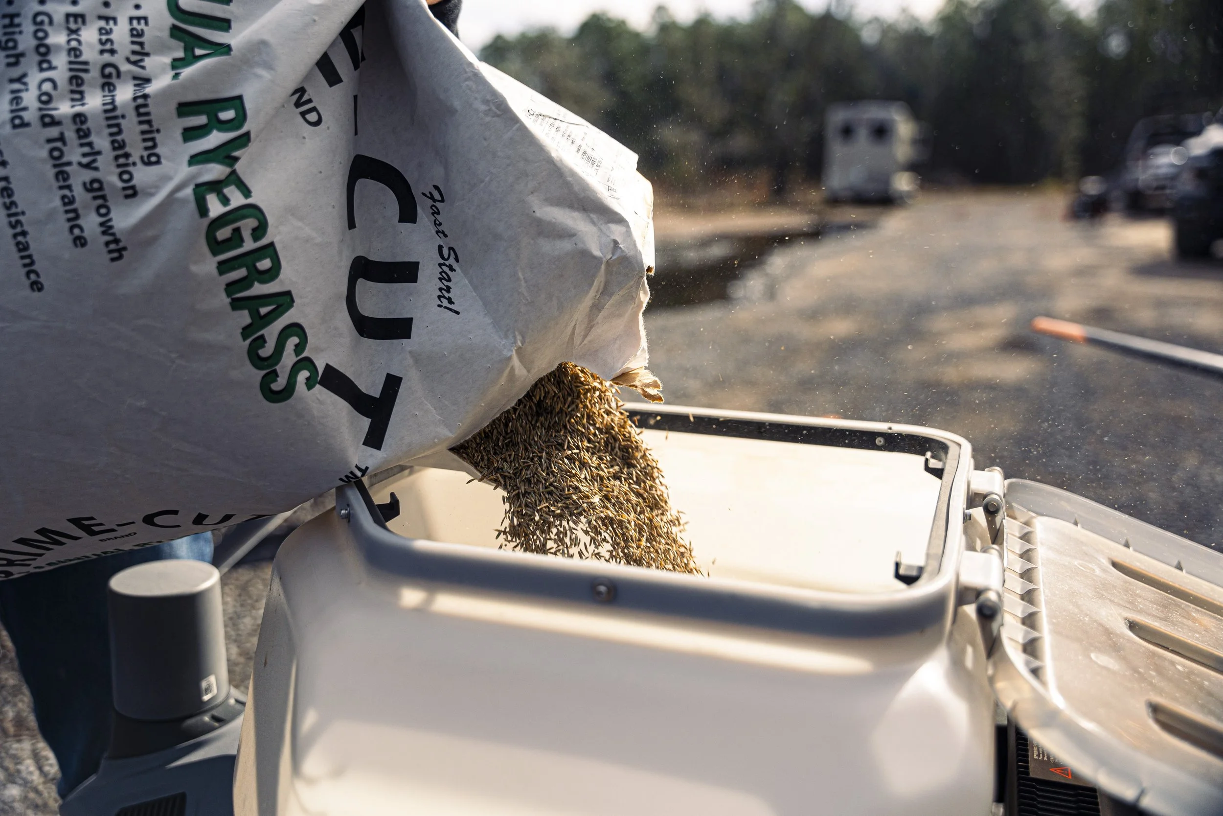 A bag of birdseed is pouring seeds into a bird feeder, with a blurred background of parked vehicles and trees.