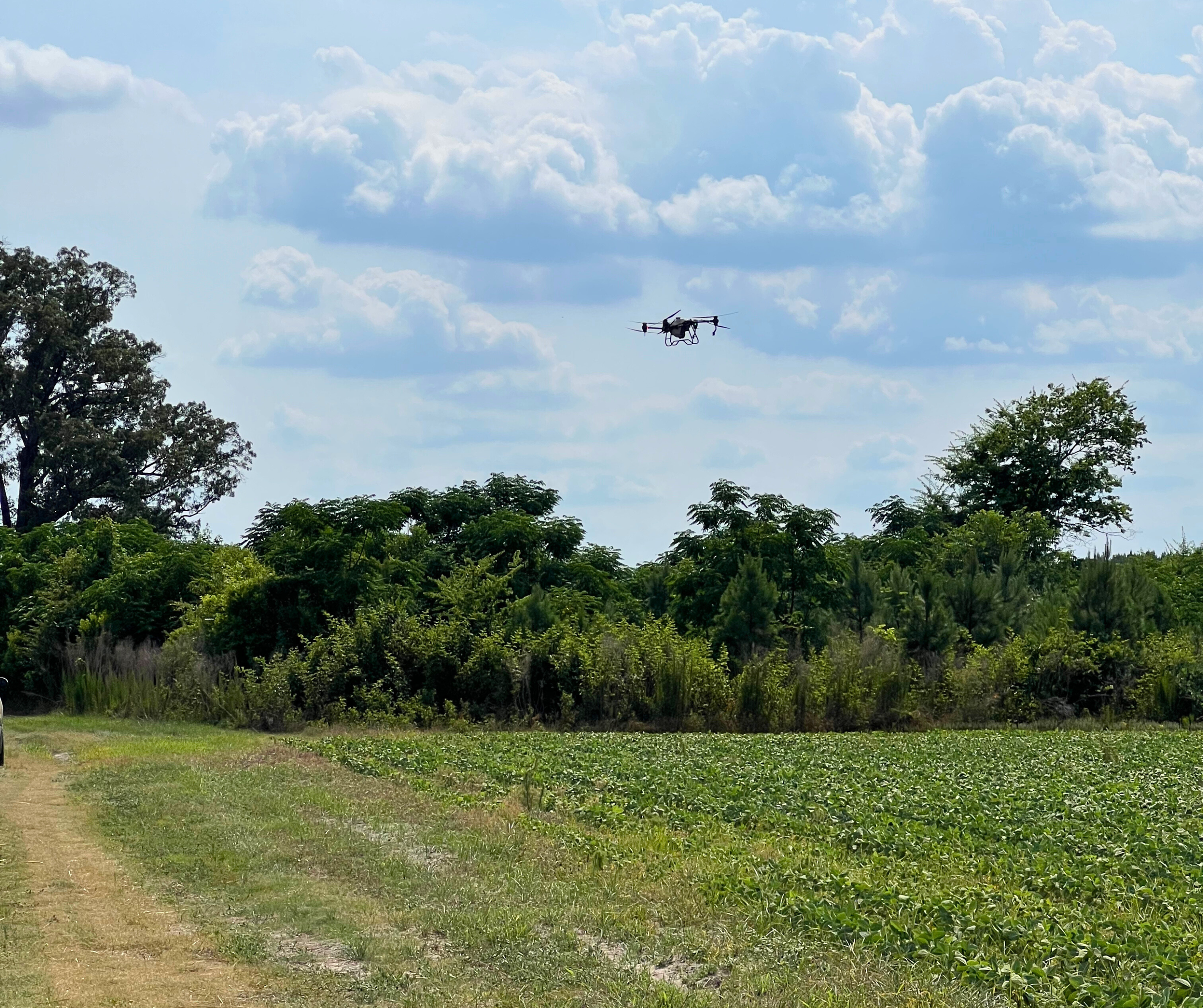 A drone flying above green trees in a rural landscape with a partly cloudy sky.
