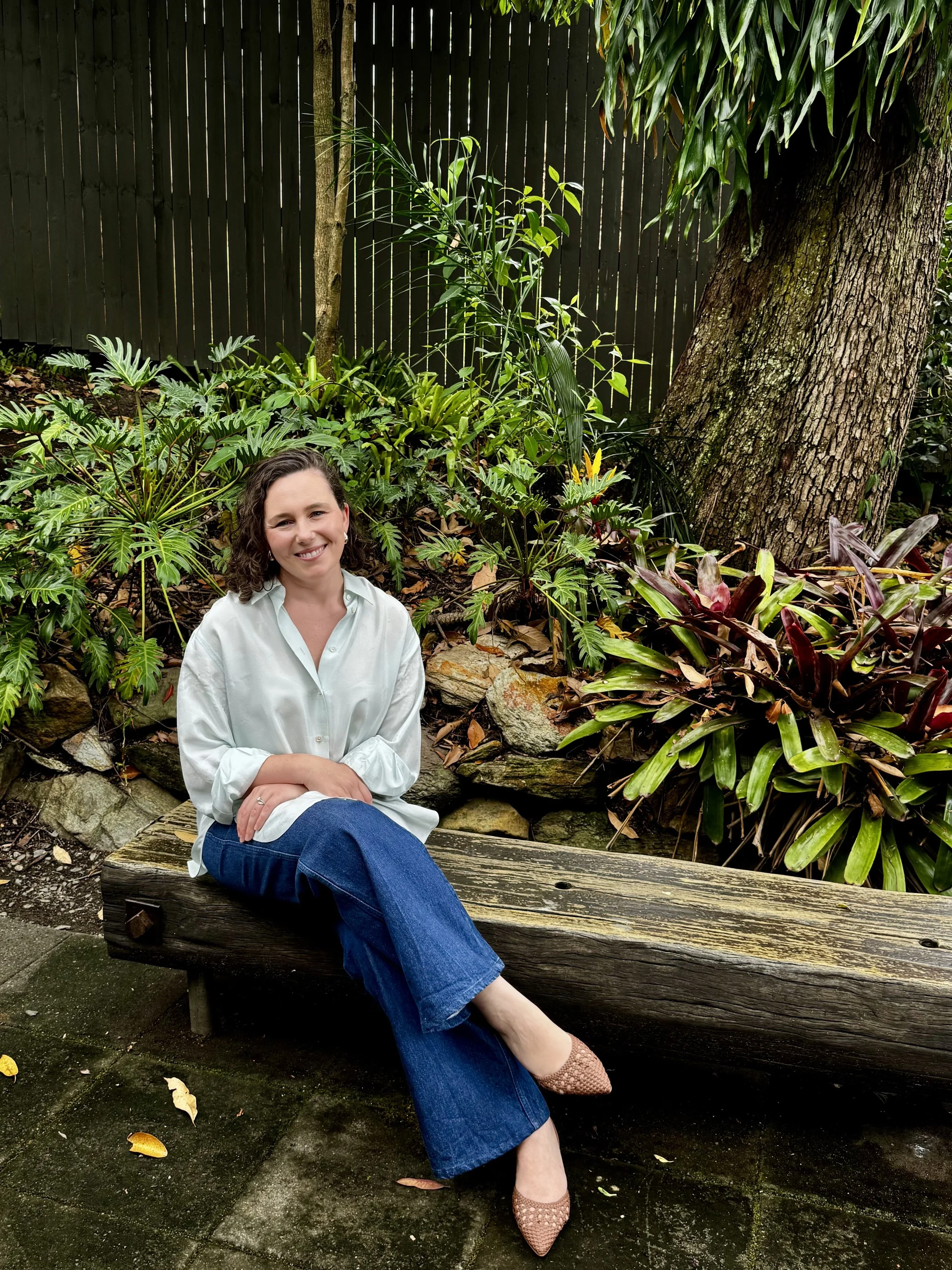 A woman with curly dark hair sitting on a wooden park bench surrounded by lush green plants and trees, wearing a white shirt, blue jeans, and pink shoes.