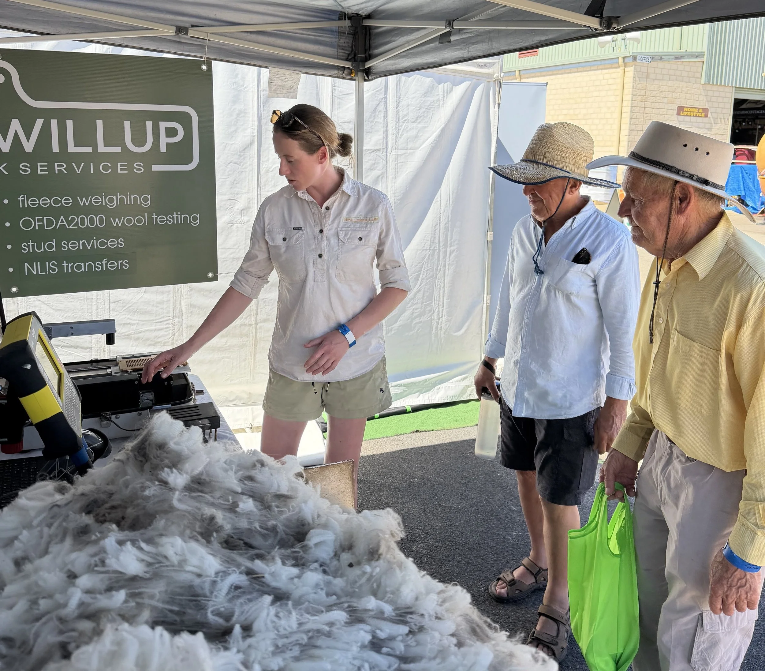Three people, two men and a woman, are at an outdoor fleece testing booth under a canopy. The woman is handling fleece samples, and the men are observing. They are wearing casual summer clothing and hats. A large green sign displays services like fleece weighing, wool testing, stud services, and NLIS transfers.