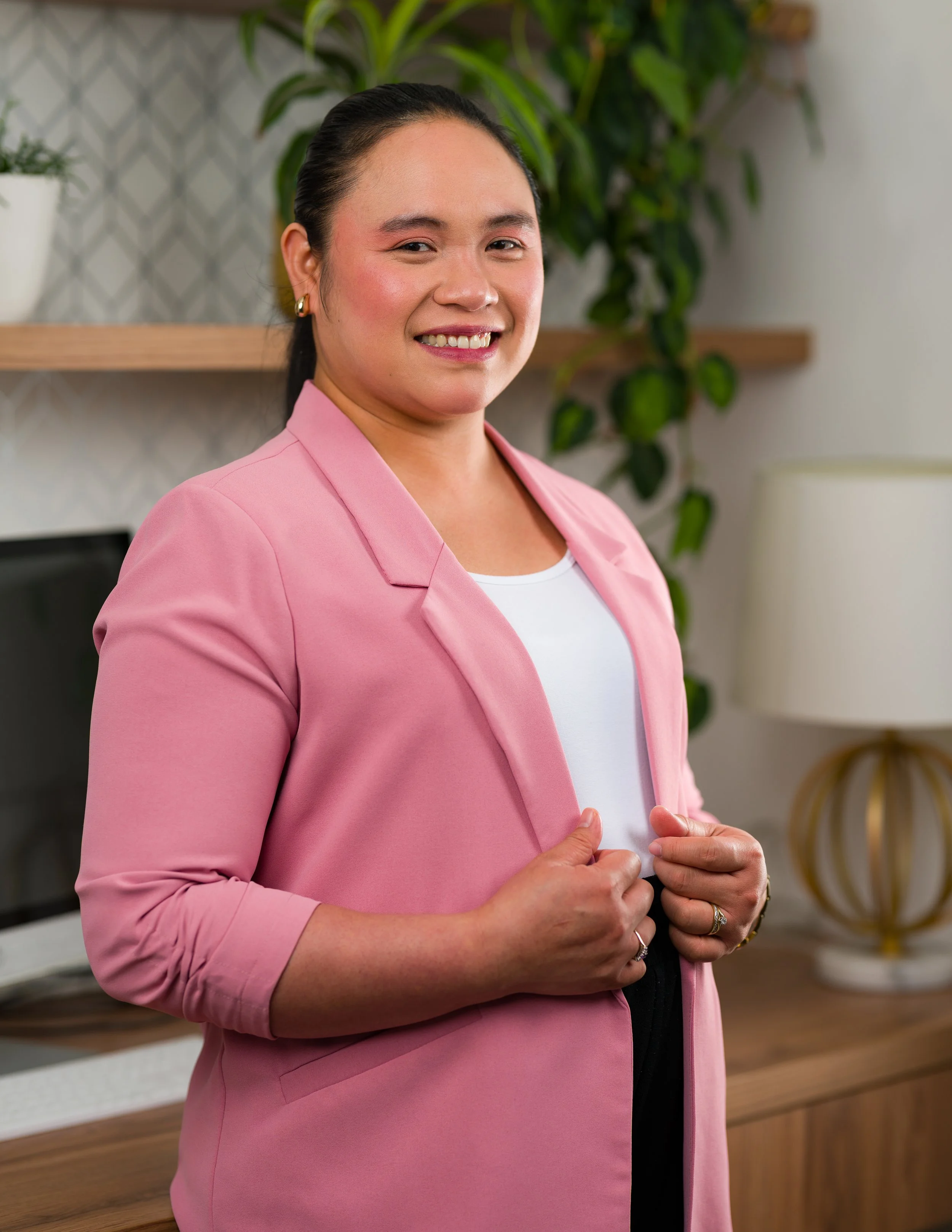 A smiling woman wearing a pink blazer and white top, standing indoors with plants and modern decor in the background.