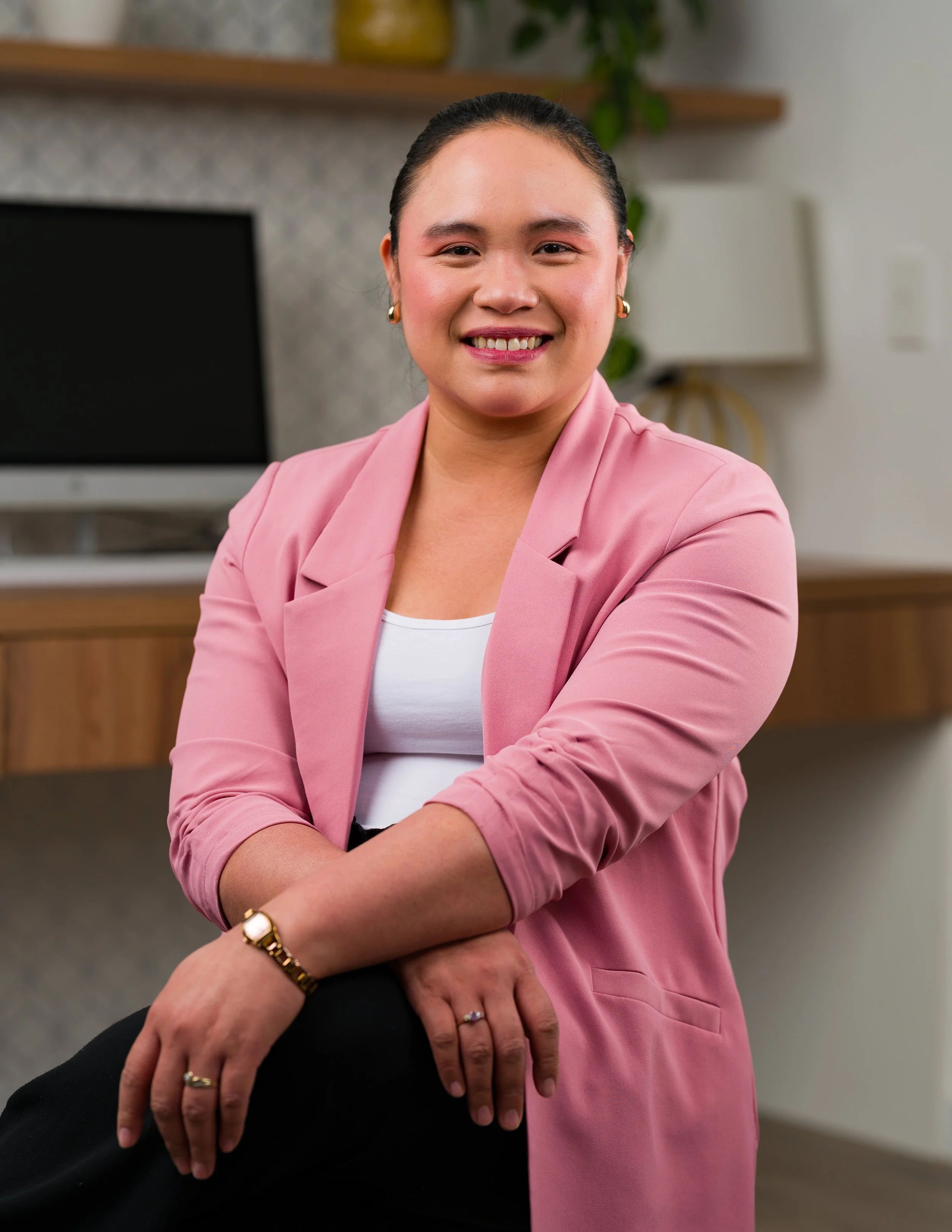 A woman in a pink blazer smiling with arms crossed, sitting in a modern office space with a computer monitor and plants in the background.