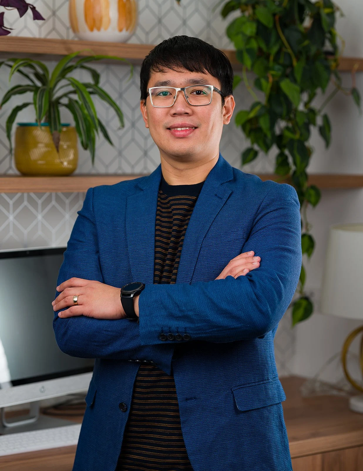 A portrait of a man with glasses in a blue blazer, standing with arms crossed in an office or home setting with potted plants and a decorative vase in the background.