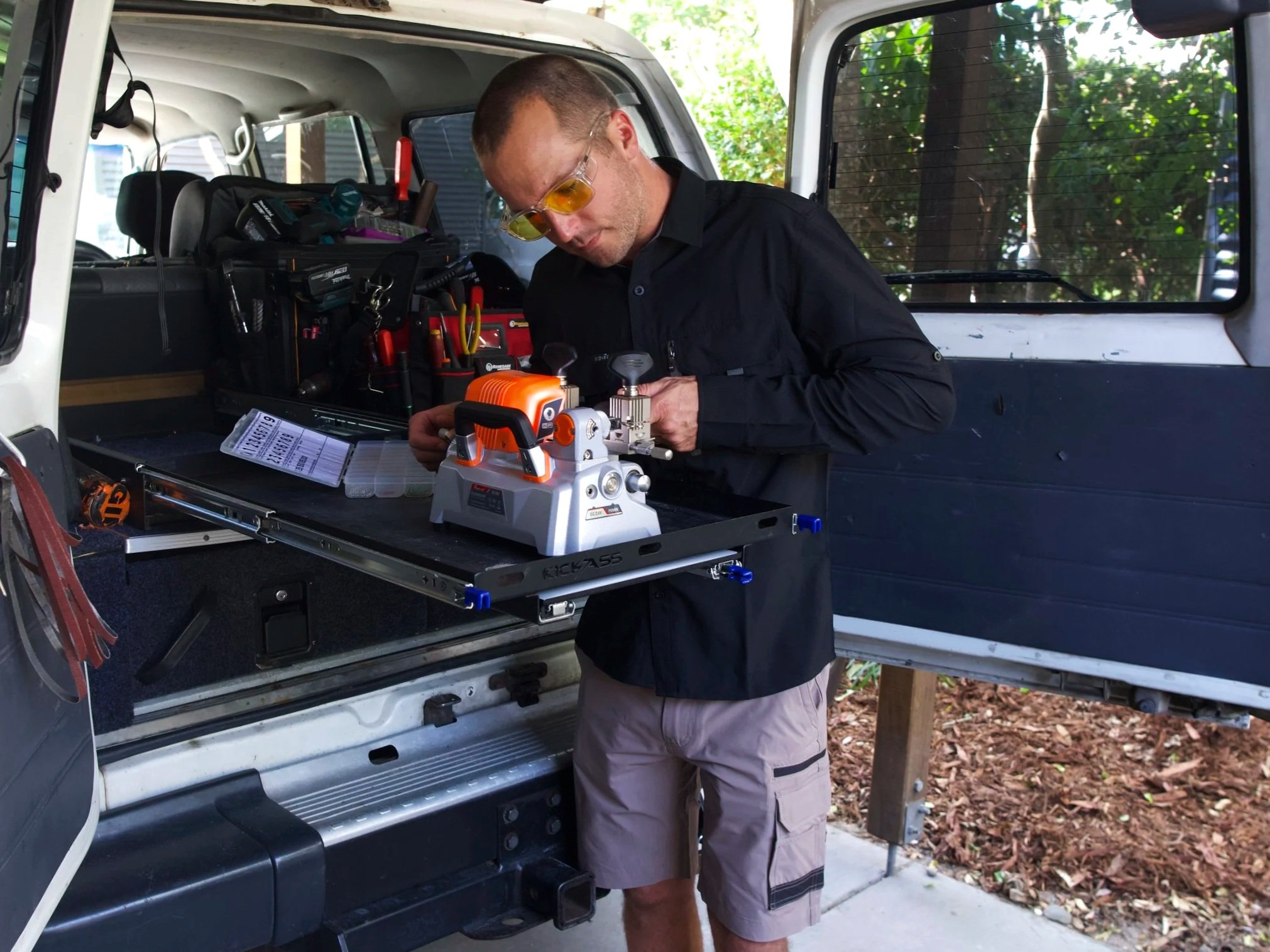 A man wearing sunglasses and casual clothes working on equipment inside the open trunk of a white SUV. The trunk contains various tools and a workbench, with trees and a house in the background.