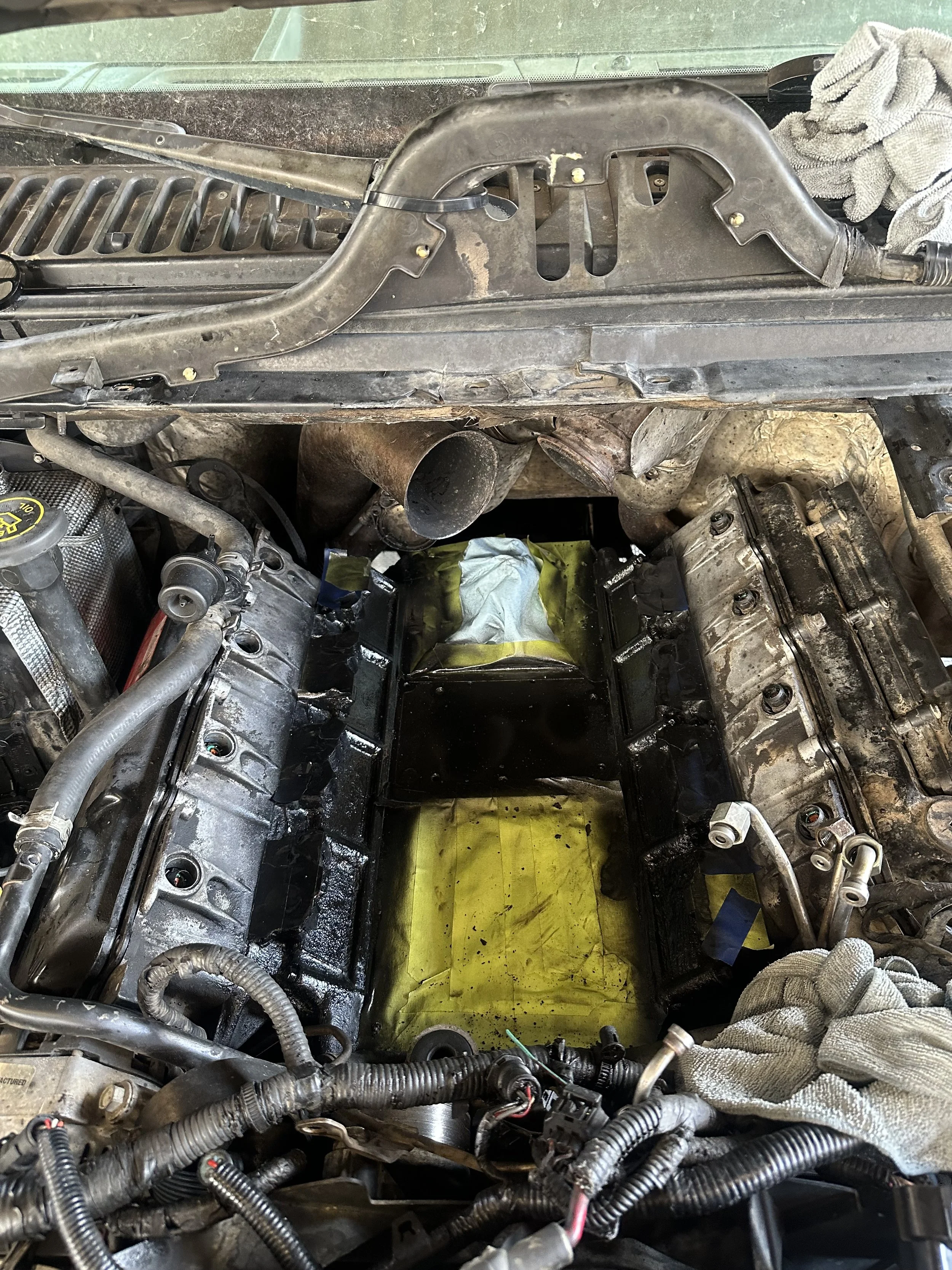Close-up of a car engine bay during repairs, showing wiring, hoses, and engine components with a yellow protective sheet underneath.