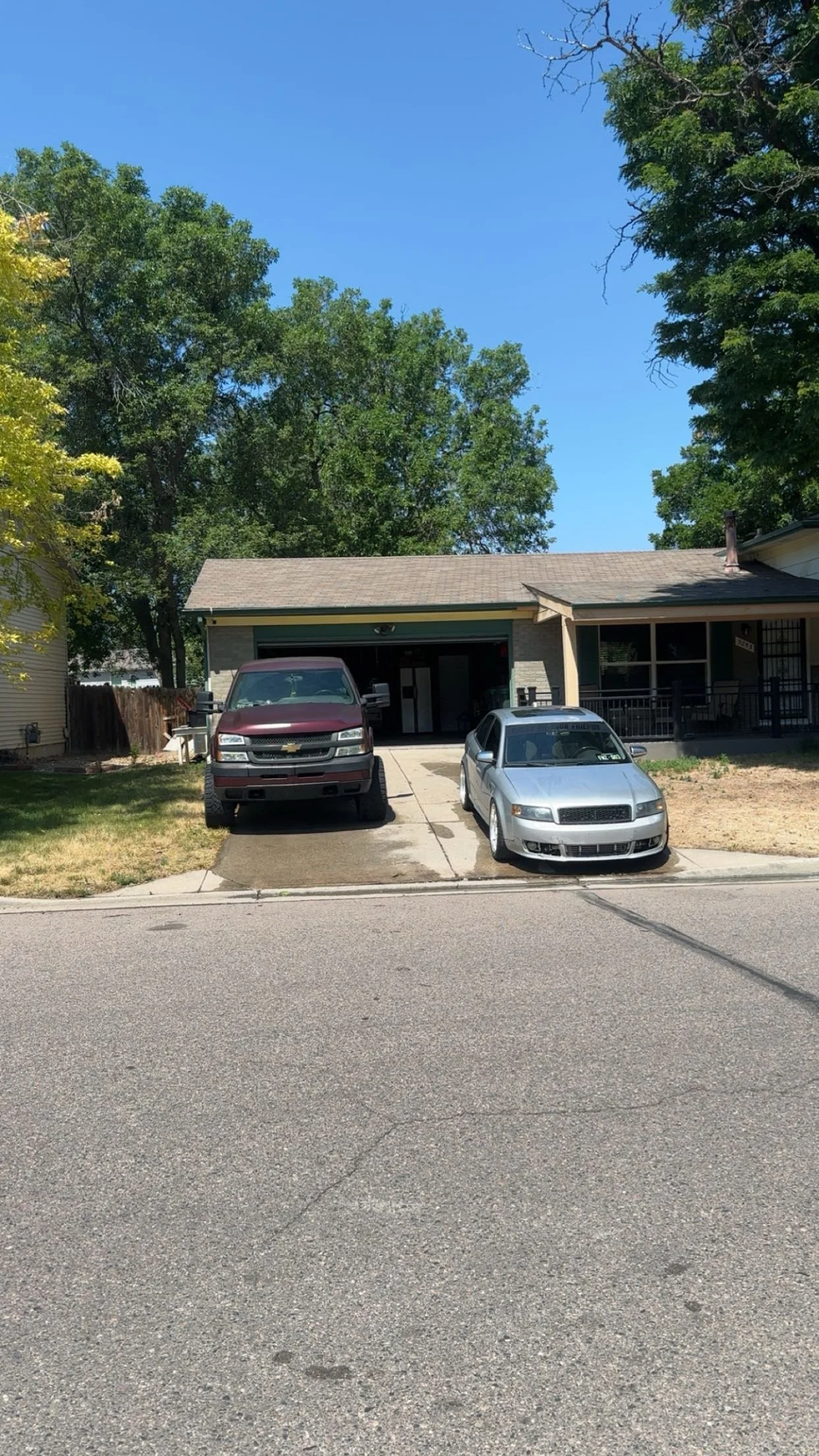 A house with a garage, two parked cars in the driveway, and trees in the background on a sunny day.