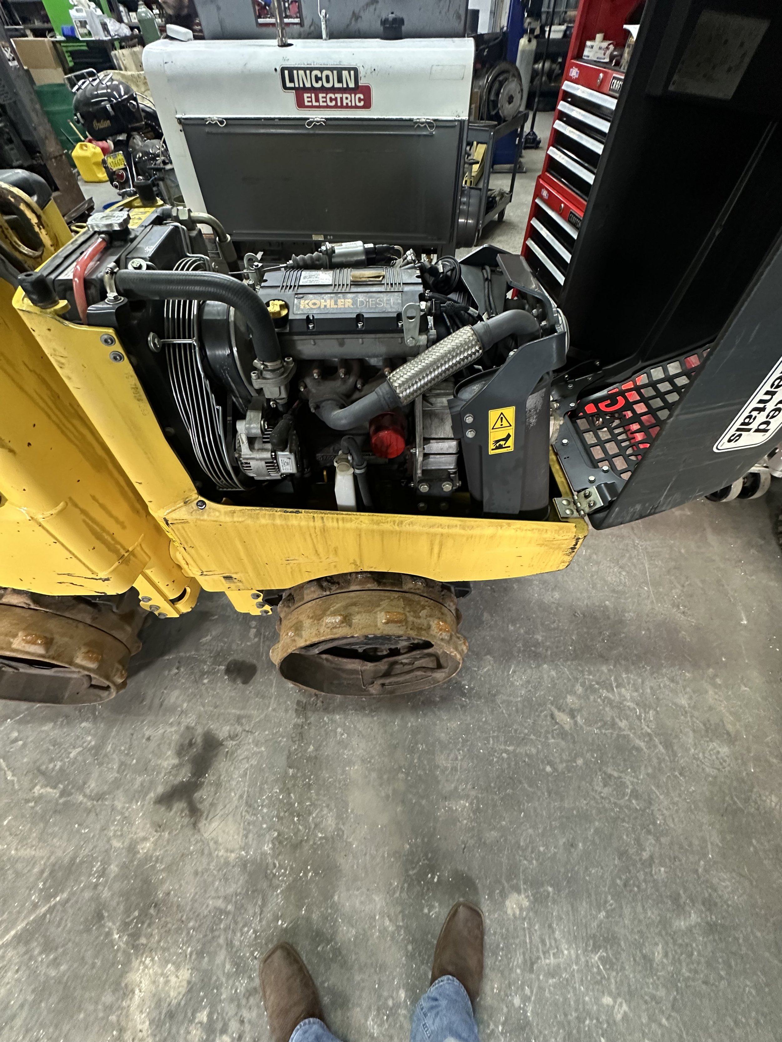View of a workshop or garage with a yellow construction vehicle, possibly a skid steer, showing the engine bay and tires, and a person's feet in brown boots and jeans standing on a concrete floor.