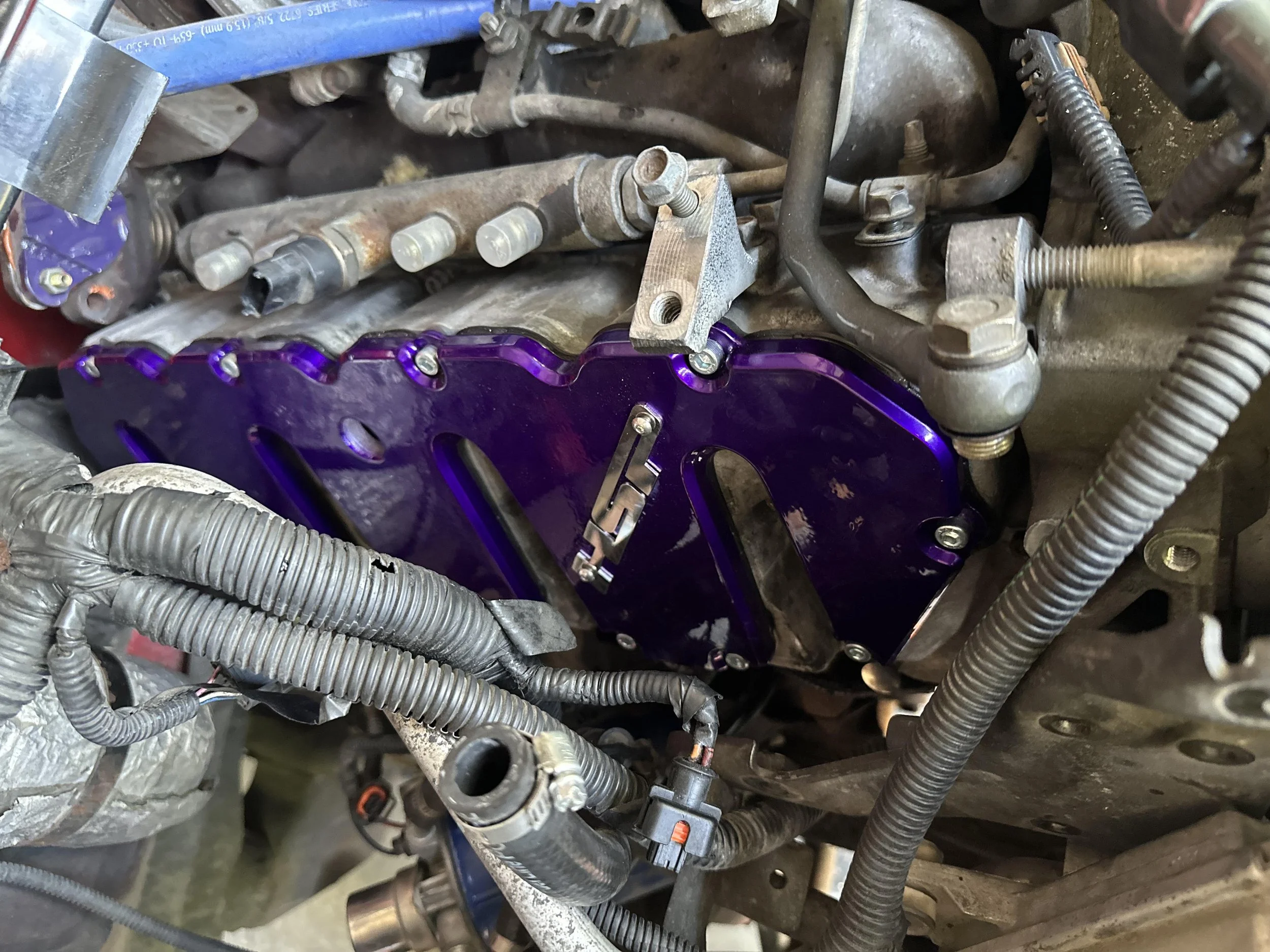 Close-up view of an engine with various metal pipes, hoses, and a purple anodized metal component. The engine parts appear used and somewhat rusty, with wires and bolts visible around the components.