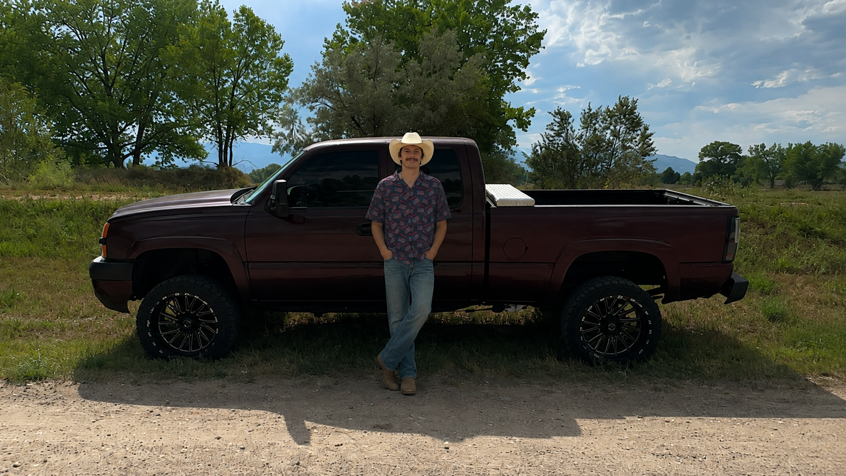 A man in a cowboy hat and patterned shirt standing in front of a maroon pickup truck on a dirt road, with trees and blue sky in the background.