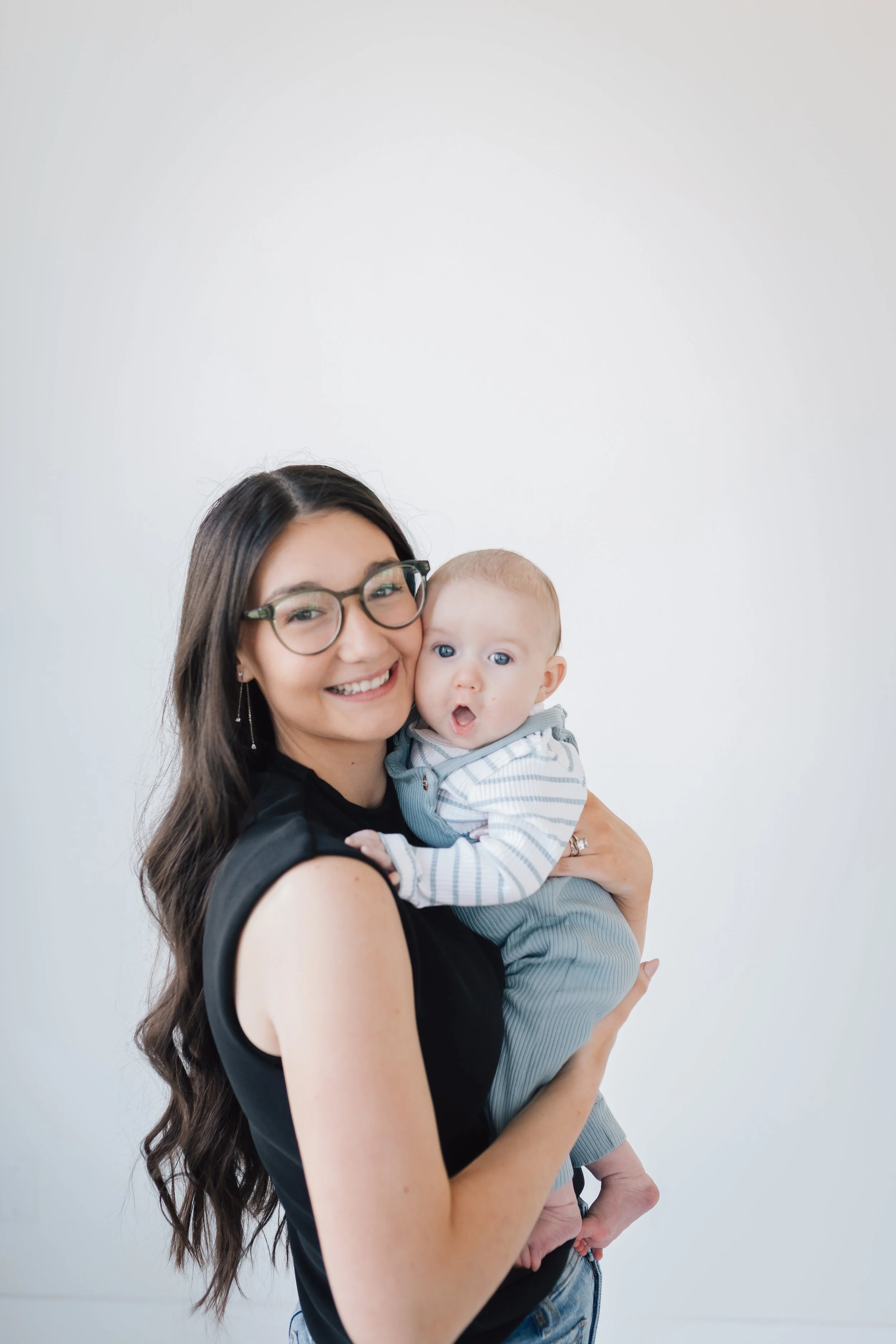 A woman with long dark hair, wearing glasses and a sleeveless black top, smiling while holding a baby with light hair and blue eyes, dressed in a striped hoodie and gray pants, against a plain white background.