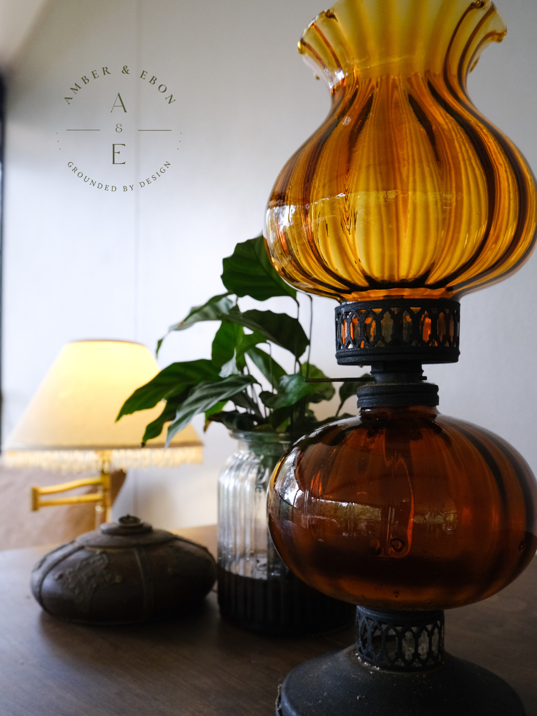 Close-up of a vintage amber glass lantern on a wooden table, with a background of a lamp and a plant in a glass vase.