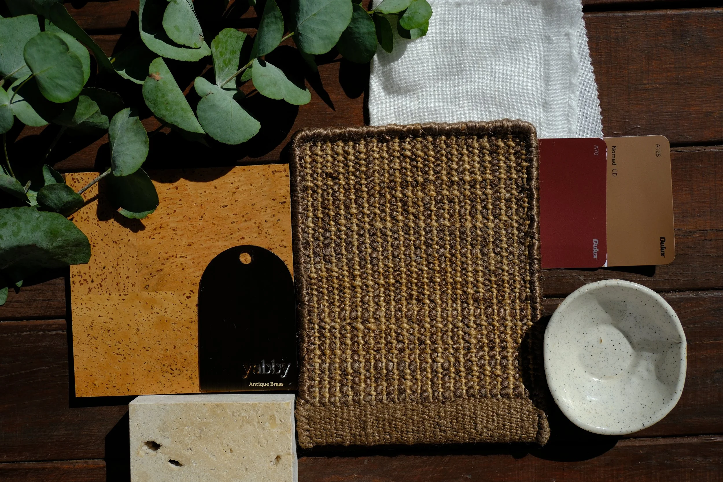 A top view of various material samples including green leaves, a white fabric, a woven brown material, a yellow textured tile, a beige stone, a black sample, and two bowls on a wooden surface.