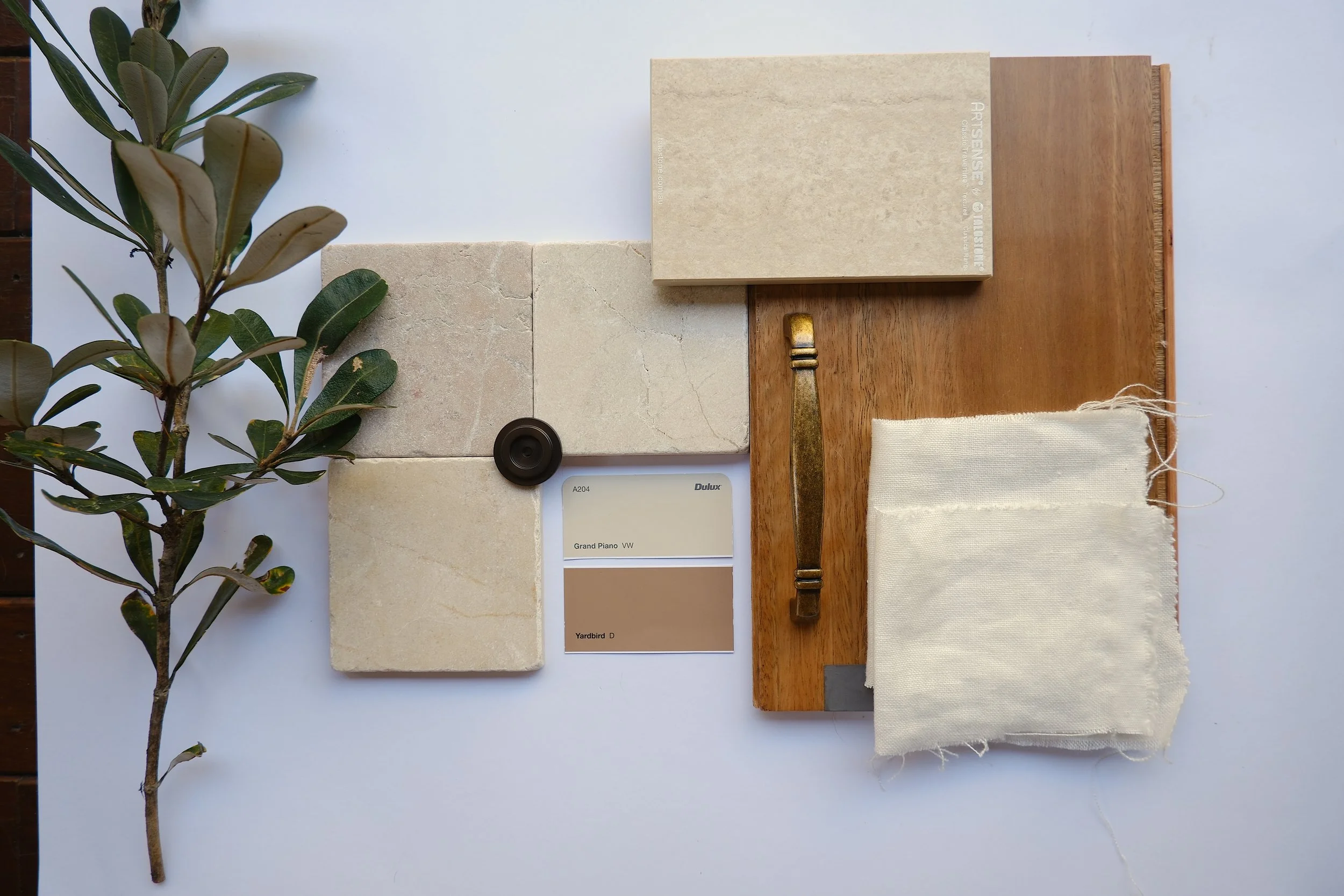 A flat lay of various interior design material samples including a plant branch, tiles, paint swatches, a book, a paint color sample, a brass hardware piece, and a fabric swatch on a white background.