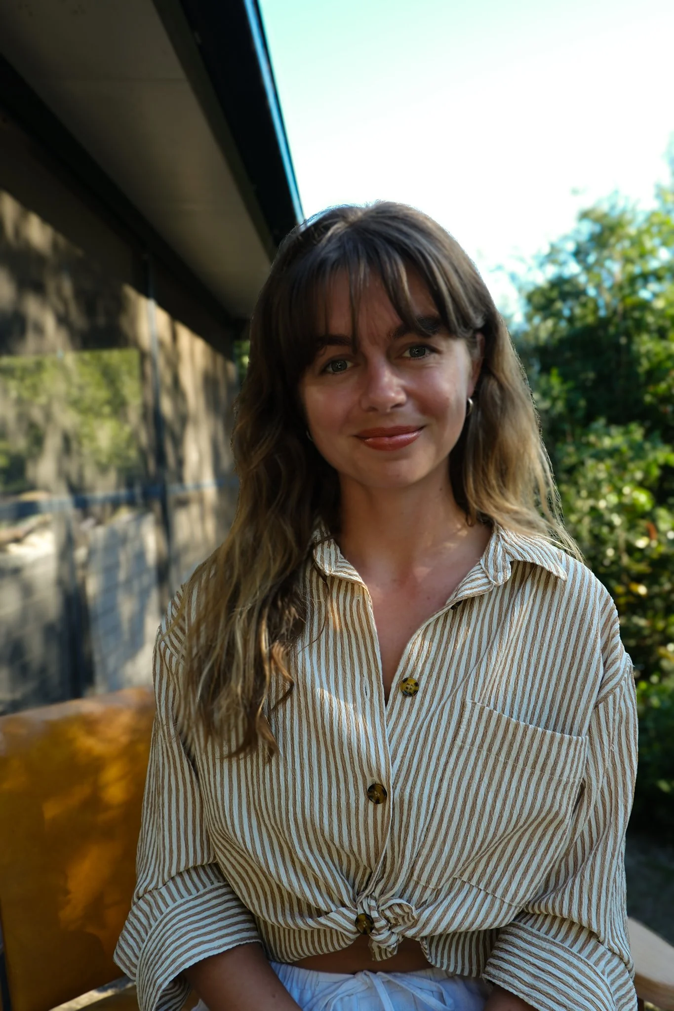 A woman with long brown hair and blue eyes sitting outdoors on a sunny day. She is wearing a beige and white striped button-up shirt and smiling softly. There are trees and a stone wall in the background.