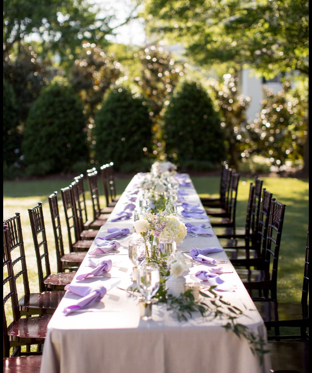 Outdoor wedding reception table decorated with flowers and lavender napkins, set in a lush garden with trees in the background.