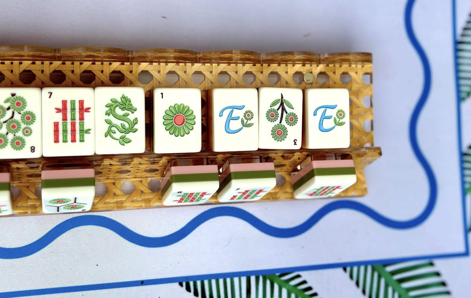 Mahjong tiles arranged to spell the word "FETE" with decorative floral patterns, placed on a plastic tray with a woven design, against a white surface with blue border lines.