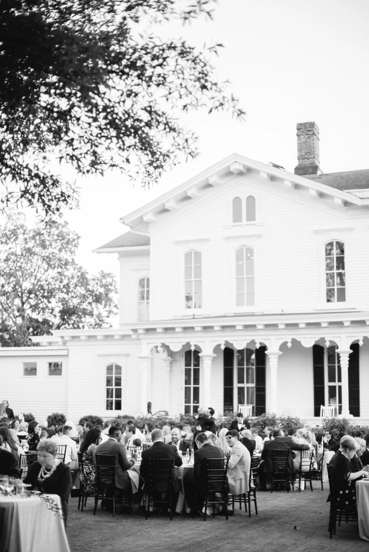 A large group of people attending an outdoor event or gathering in front of a historic white house with columns and tall windows, surrounded by trees.