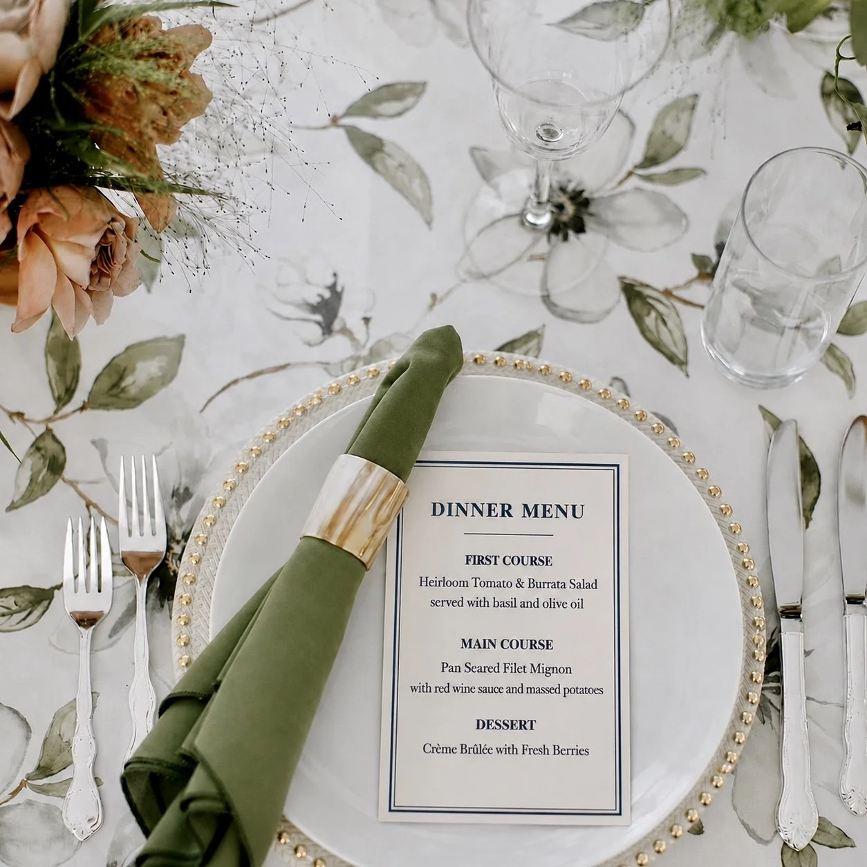 Elegant table setting with a dinner menu, green napkin with gold napkin ring, crystal glasses, and floral centerpiece on a leafy patterned tablecloth.