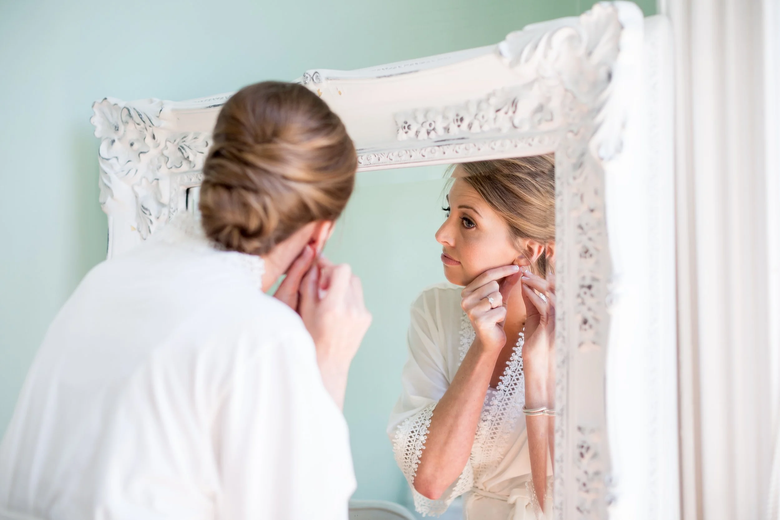 A woman with styled hair looking at herself in an ornate mirror, touching her face and wearing a white lace top.