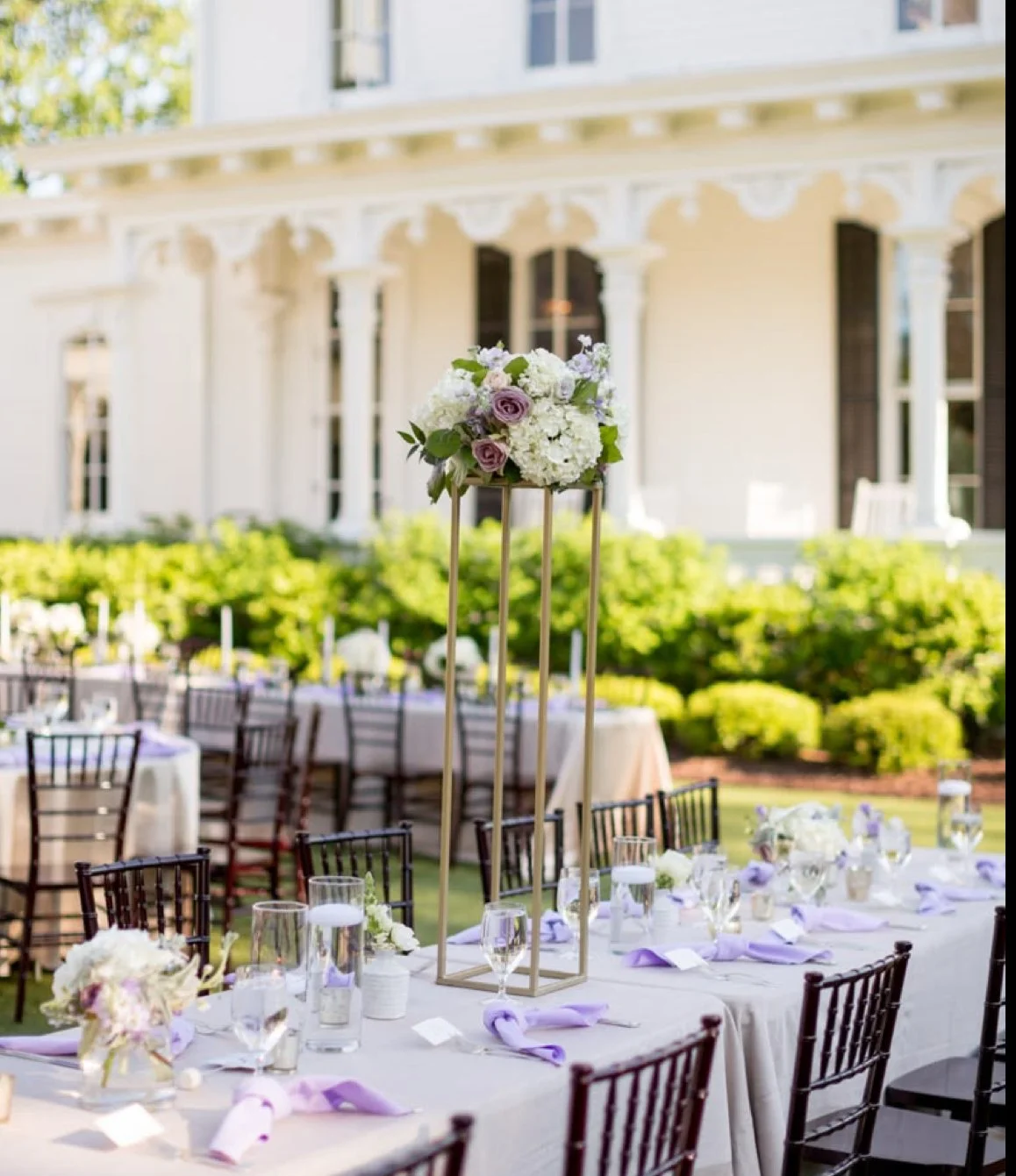Outdoor wedding reception tables with purple and white floral centerpieces and purple napkins, set in front of a white house with a porch and lush greenery.