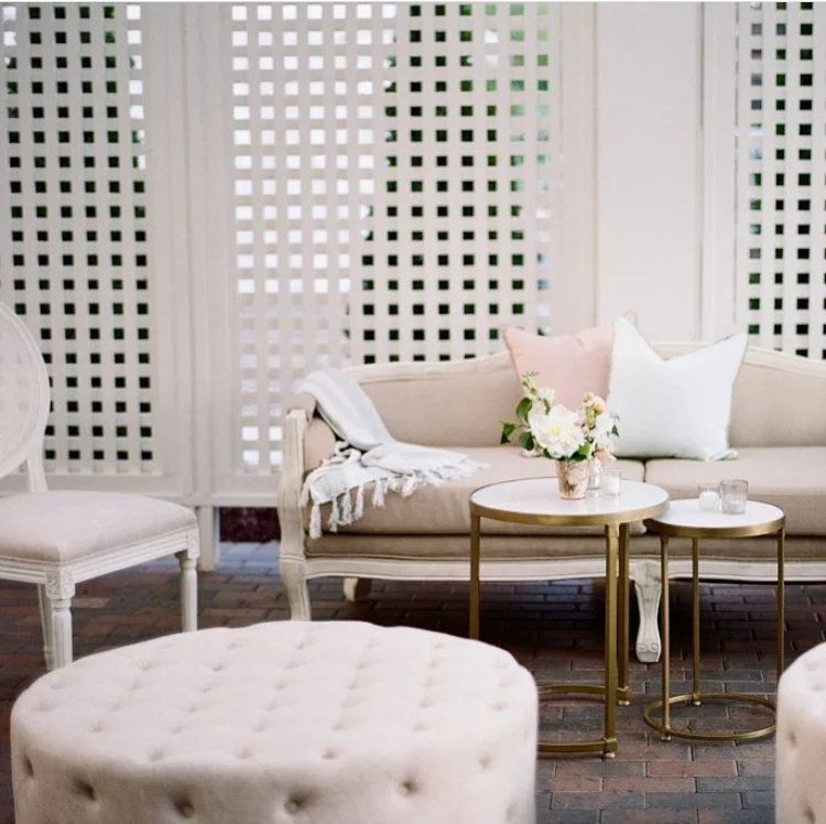 A cozy living room corner with a beige vintage sofa, pink and white throw pillows, a white blanket, and a floral arrangement on round nesting tables. White lattice panels background with brick flooring.