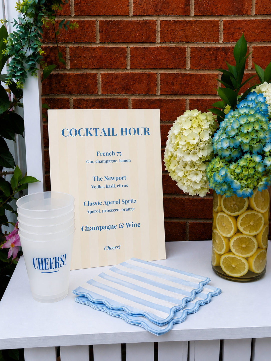 A sign listing cocktail options for a cocktail hour, including French 75, The Newport, Classic Aperol Spritz, and Champagne & Wine. The sign is on a table with striped napkins, plastic cups labeled 'Cheer!', and a flower vase with white and blue hydr