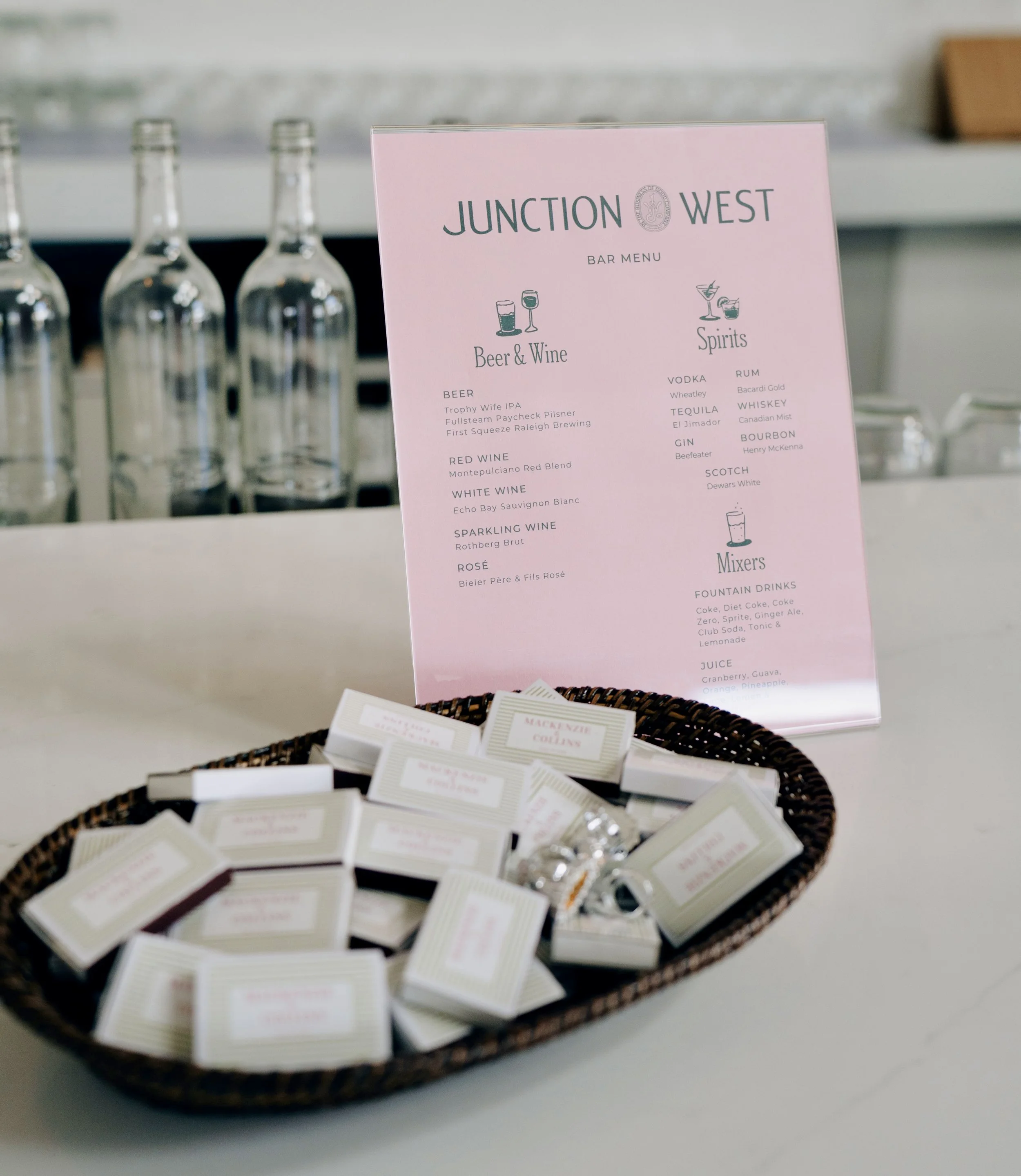 Pink bar menu sign on a table with drink options at a bar, featuring beer, wine, spirits, and mixed drinks, with bottles in the background and small packages in a basket in the foreground.