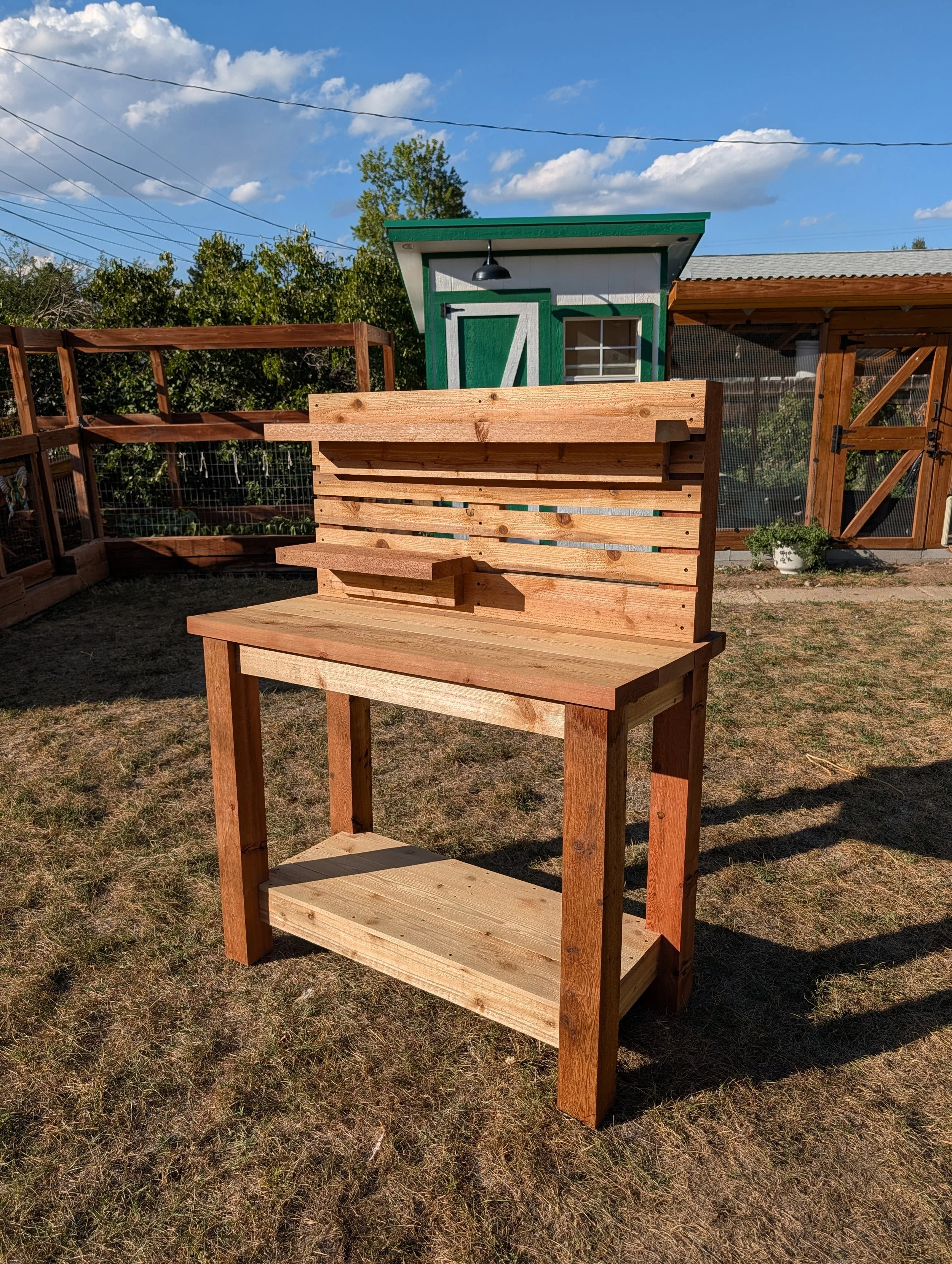Freshly built wooden outdoor shelving unit with three shelves, situated on a patch of dry grass, with a background of a small green and white shed, a fenced garden area, and a blue sky with some clouds.