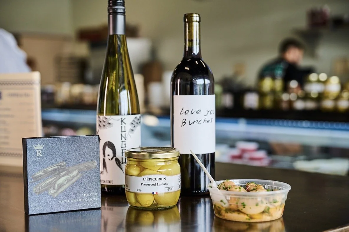 Two bottles of wine, a jar of preserved lemons, a box of smoked petit razor clams, and a container of shrimp salad on a counter in a cafe or restaurant.