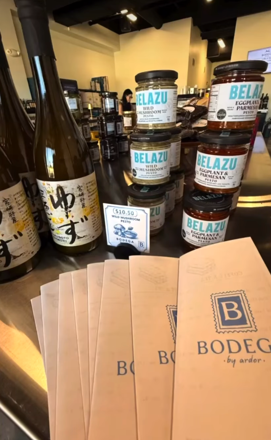 Stacked jars of BELAZU eggplant and parmesan pesto and wild mushroom pesto on a table at a store, with a sign indicating the price of $10.50 for the wild mushroom pesto, and BODEGA brochures in the foreground.