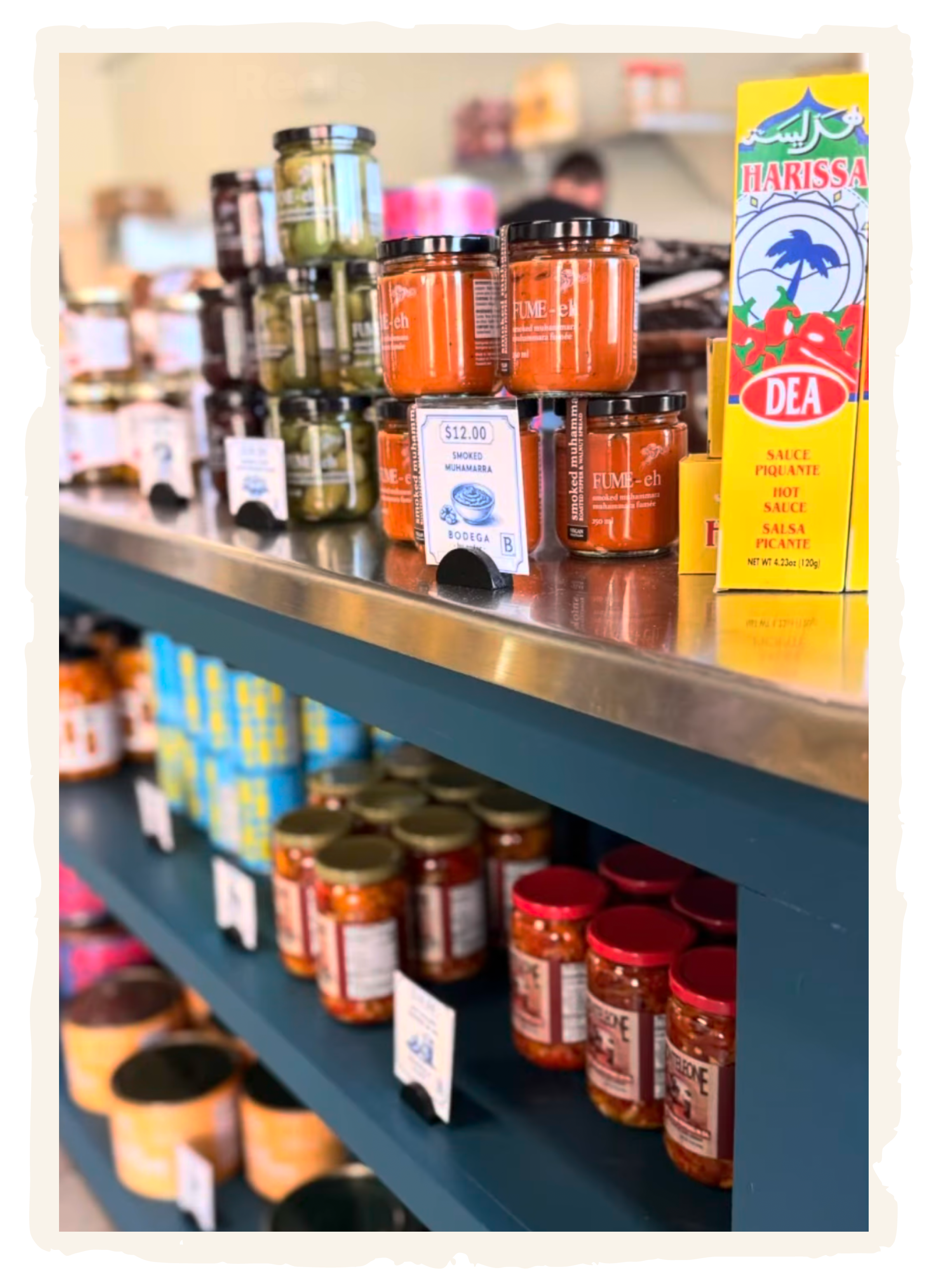 Shelf with jars of sauces and condiments, including Harissa hot sauce and smoked muhamara in a grocery store.