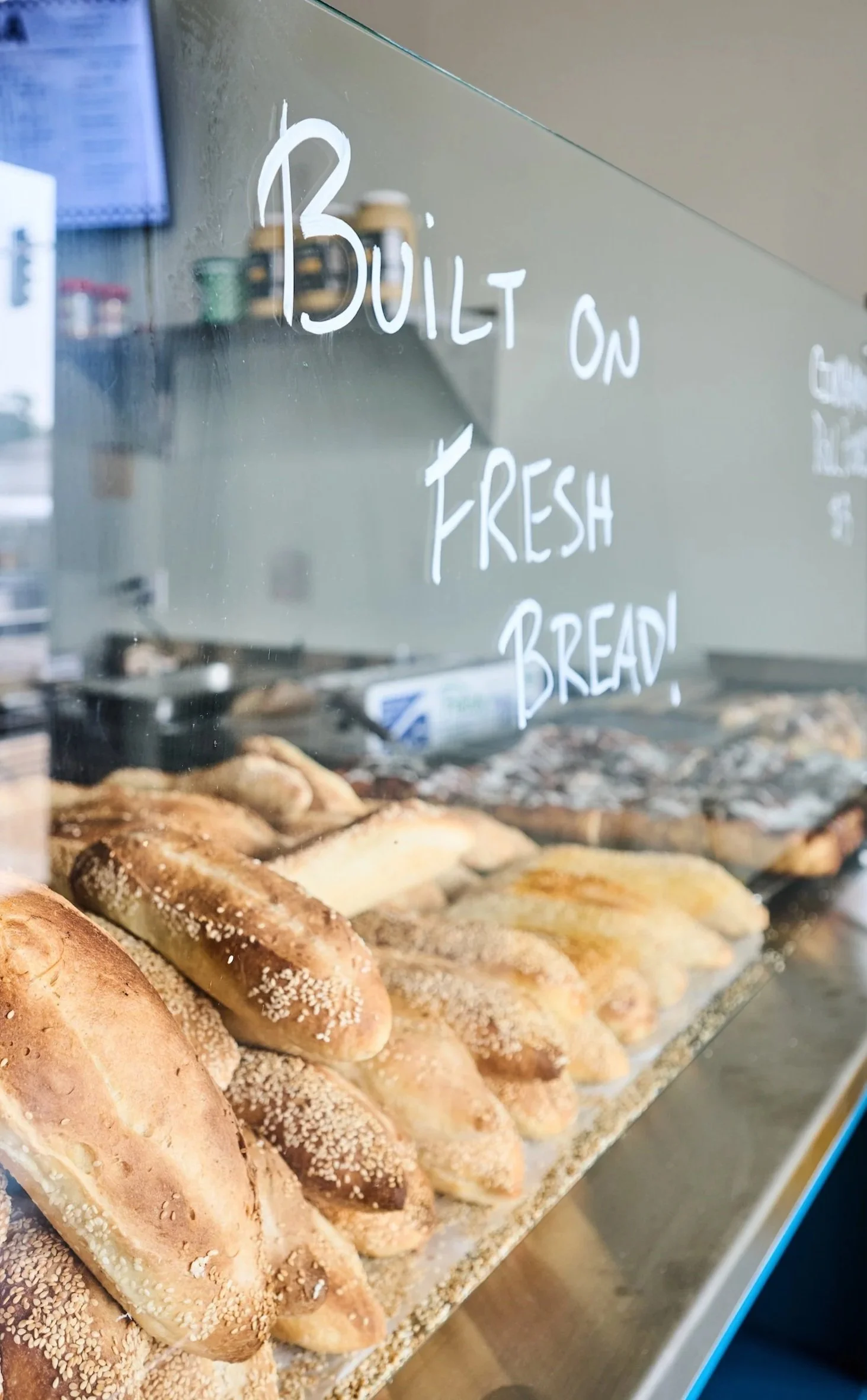 A display of assorted freshly baked bread and pastries behind a glass case with a handwritten sign saying 'BULIT ON FRESH BREAD'.