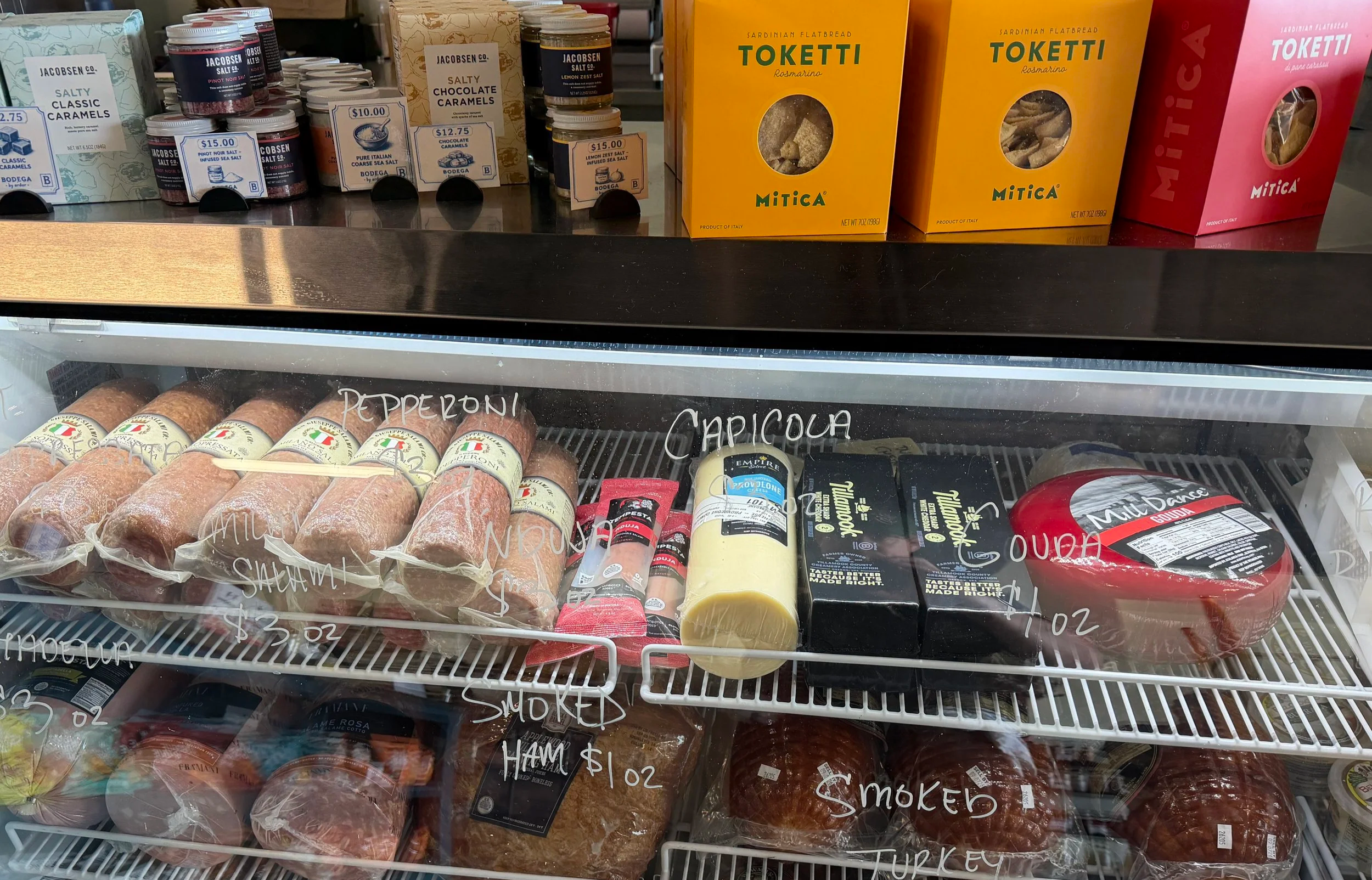 Display of assorted food items in a refrigerated case, including sausages, cheese, and packaged meats, with handwritten labels and prices on the glass.