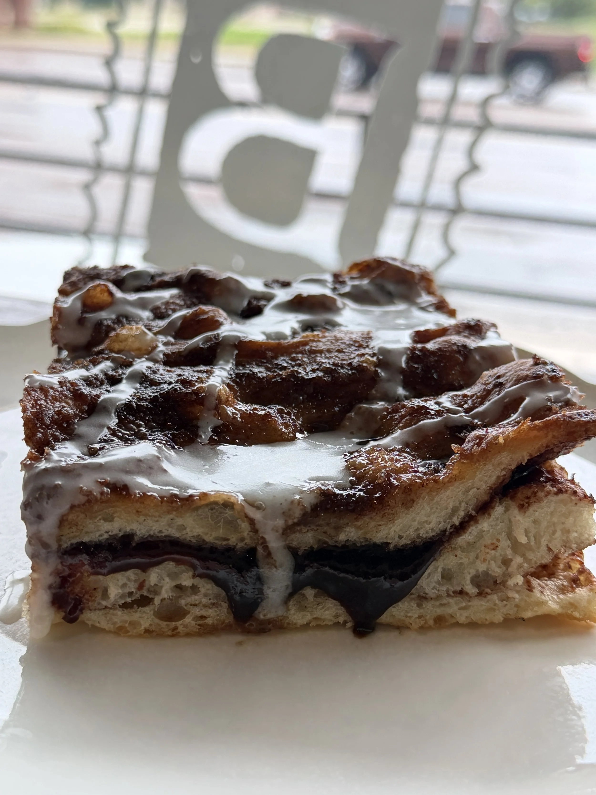 A slice of cinnamon roll cake with icing, chocolate, and nuts on top, placed on a white surface with a blurred background of a street scene and a metal decorative piece.