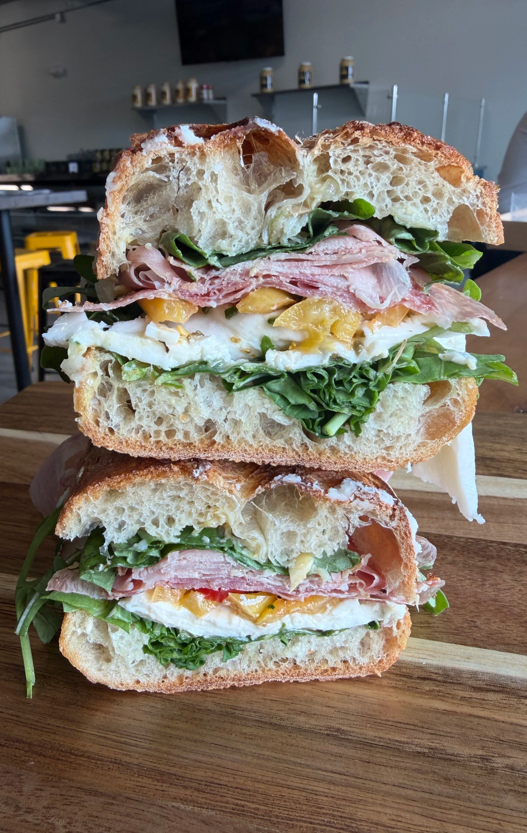 Close-up of a stacked sandwich with various meats, cheese, greens, and bread on a wooden board in a restaurant setting.