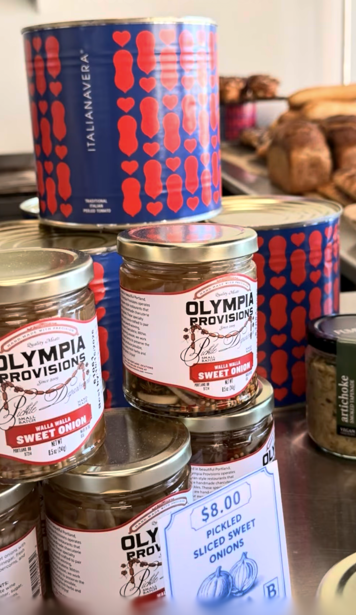 Cans of Olympia Provisions pickled sliced sweet onions on store shelves, with a sign indicating $8.00 for pickled sliced sweet onions.