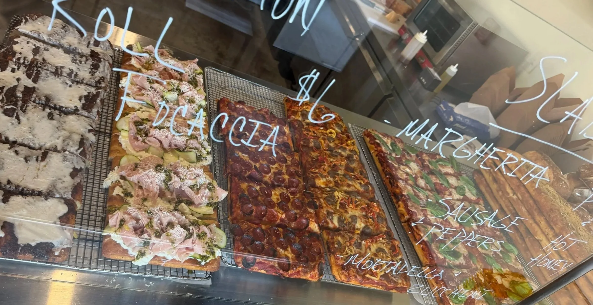 Display case with various types of pizza and baked goods, some with toppings like pepperoni, vegetables, and cheese, seen through a glass window with handwritten menu items on the glass.