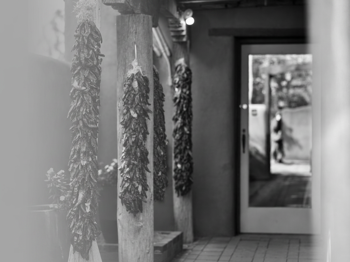 Black and white photo of dried chili peppers hanging on vertical wooden poles near a door with glass window, leading outside where a person is walking.