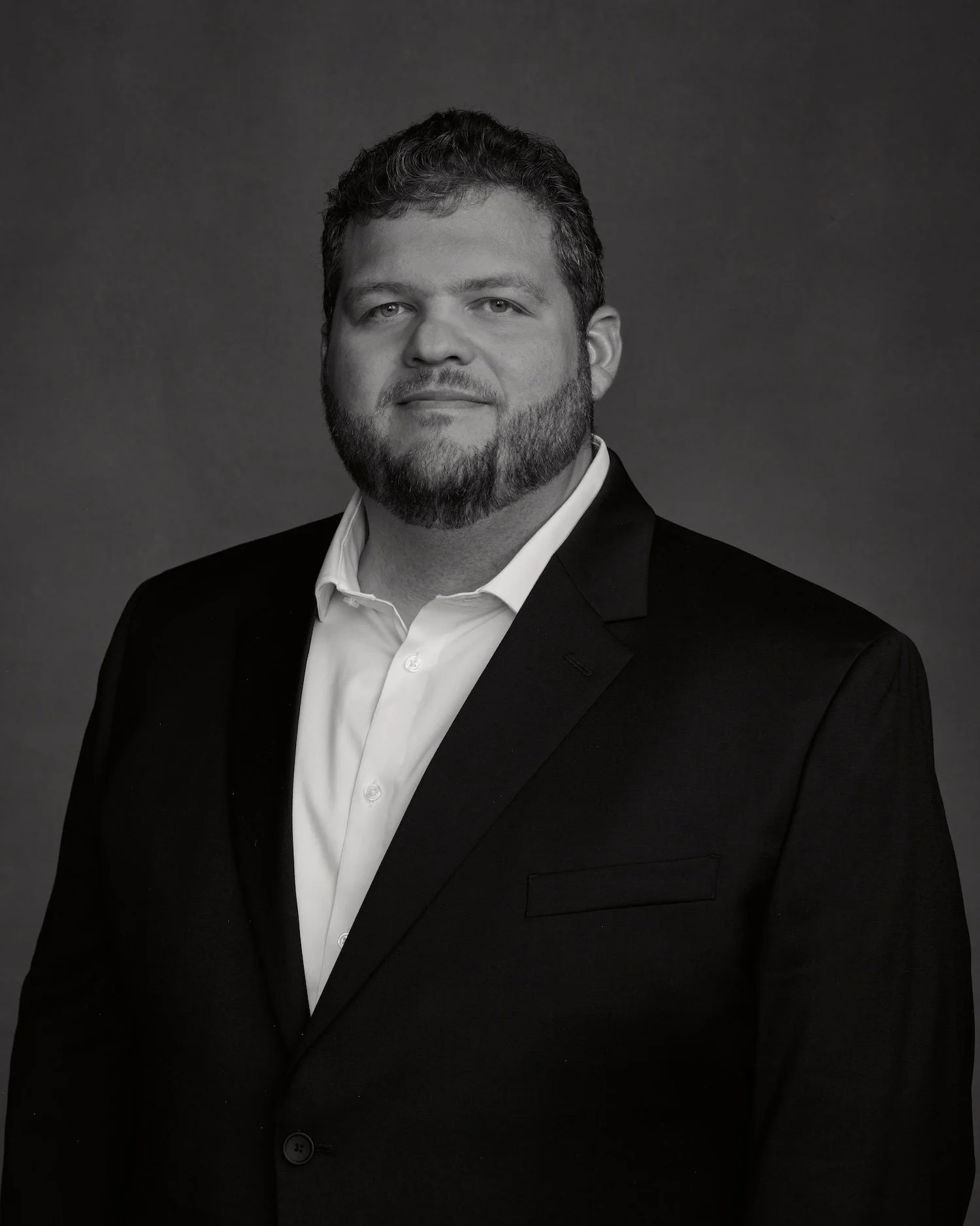 Black and white portrait of a man in a suit and white shirt, with short dark hair and a beard, looking into the camera against a plain background.