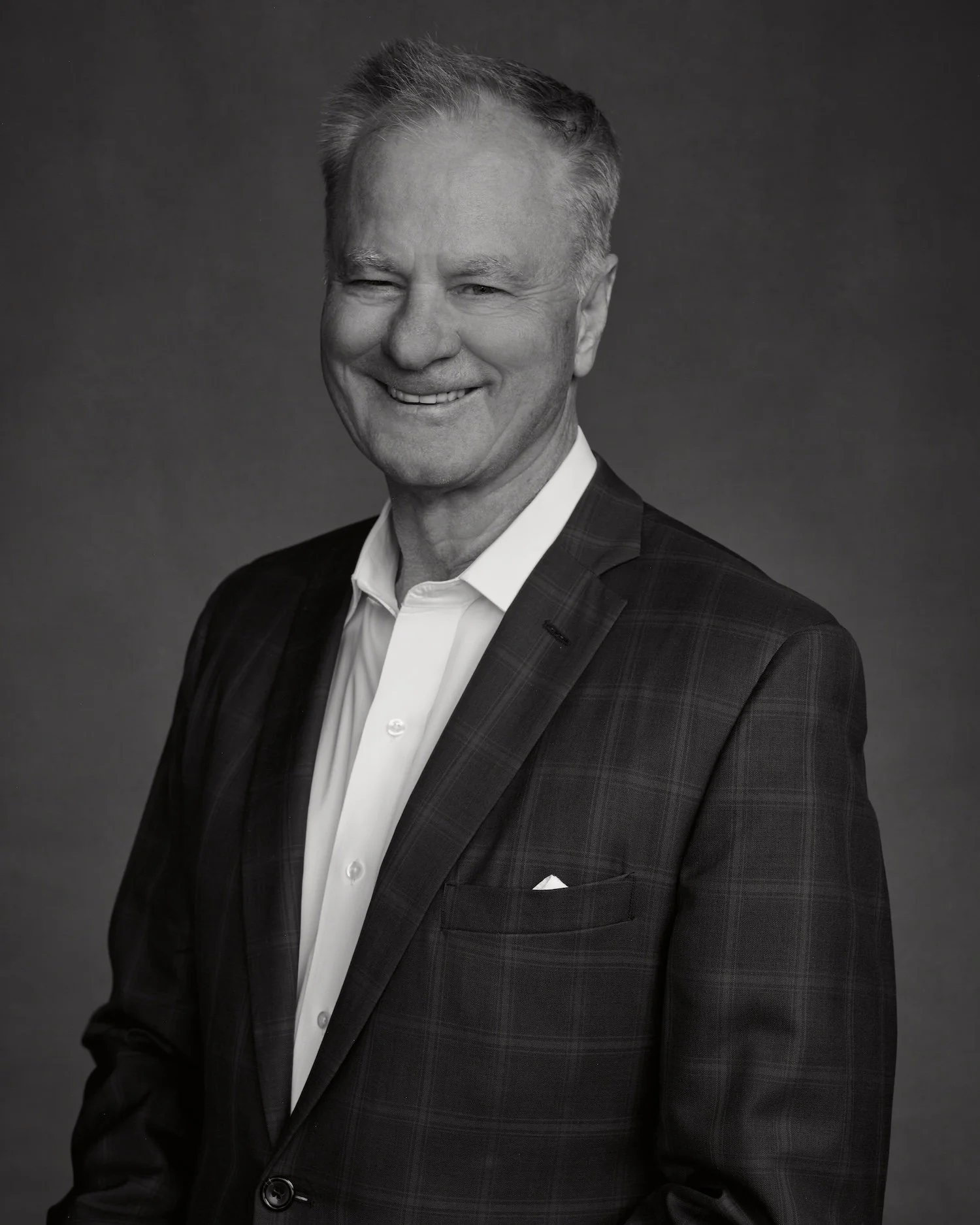 A black and white portrait of a smiling middle-aged man in a suit, winking with one eye, against a dark background.