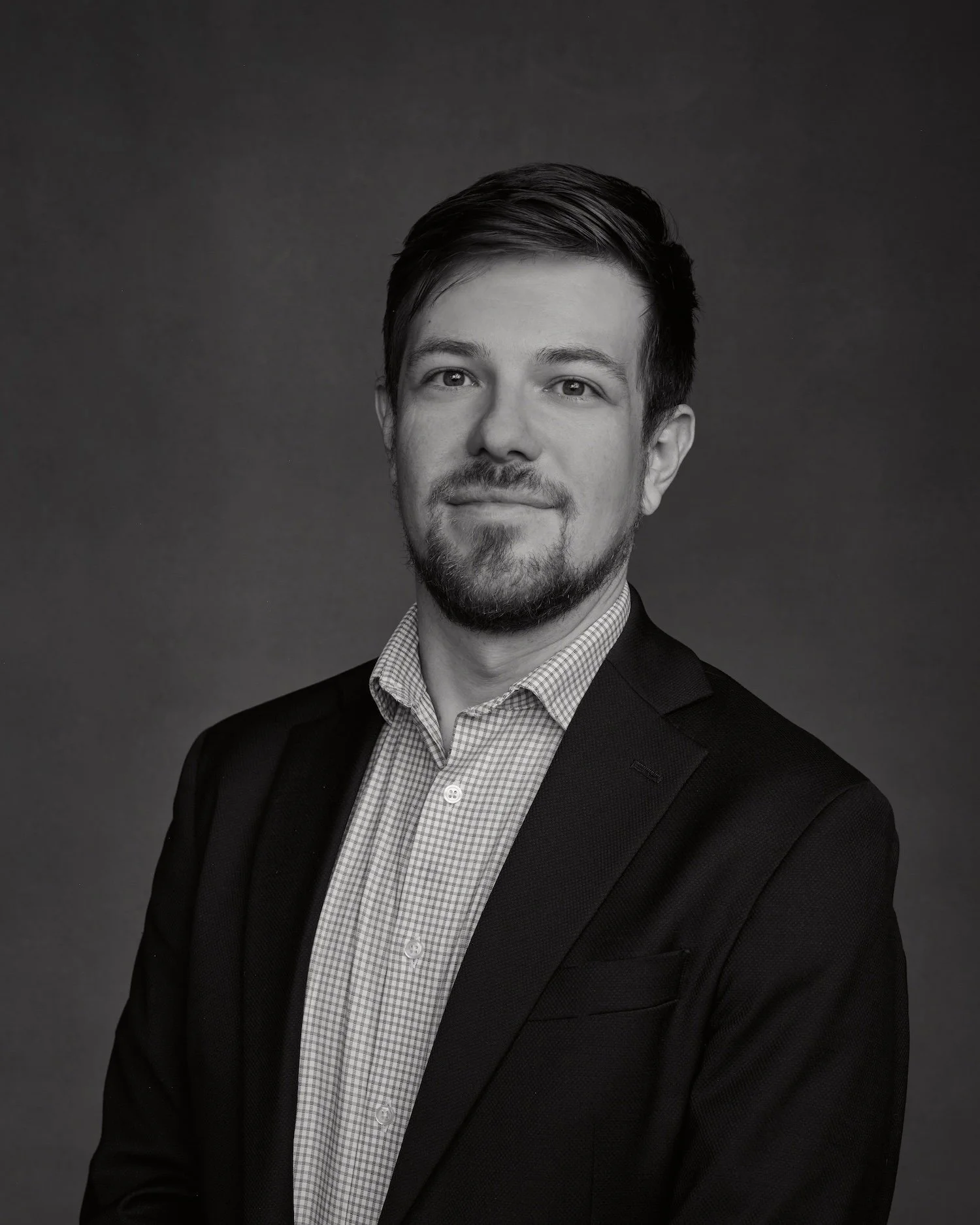 Black and white portrait of a man with short, dark hair and a beard, wearing a suit jacket and light-colored shirt, looking at the camera against a dark background.