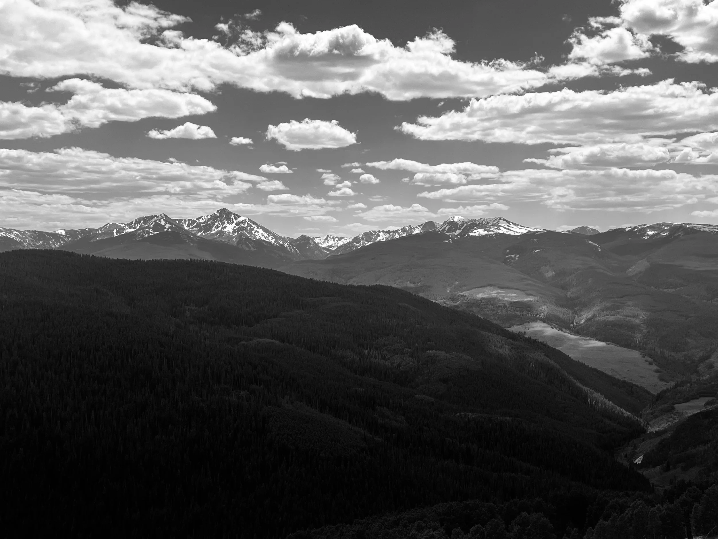 Black and white photo of mountains with snow on their peaks, layered under a sky filled with scattered clouds.