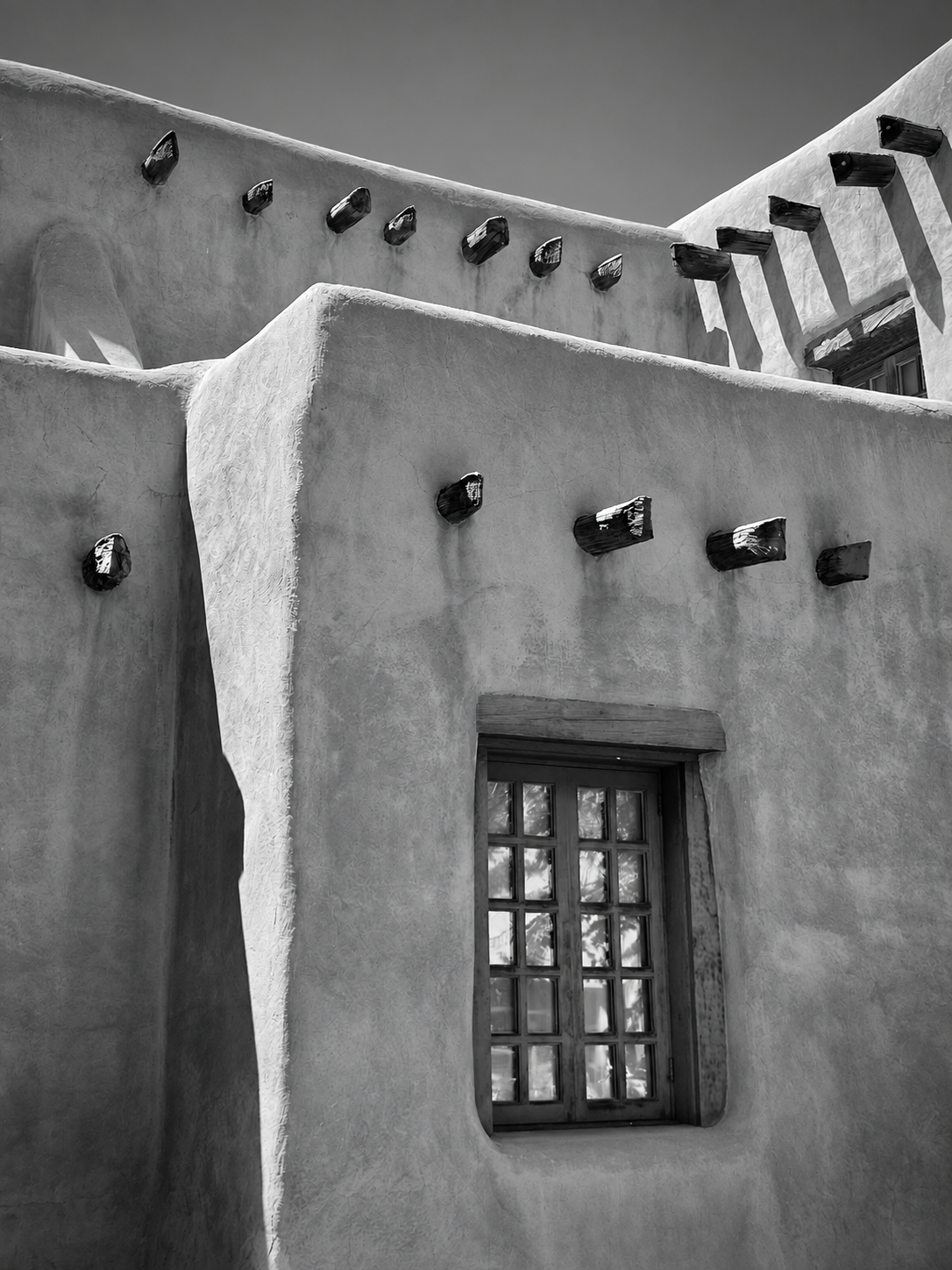 A close-up of traditional adobe building with wooden beams projecting from the walls and a wooden window with small panes, in black and white.
