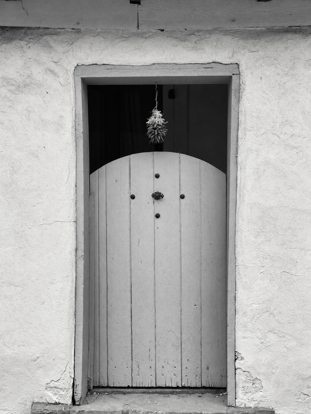 A wooden door with decorative black knobs and a handle, set in a textured wall, with a small window above the door holding a hanging pinecone decoration.