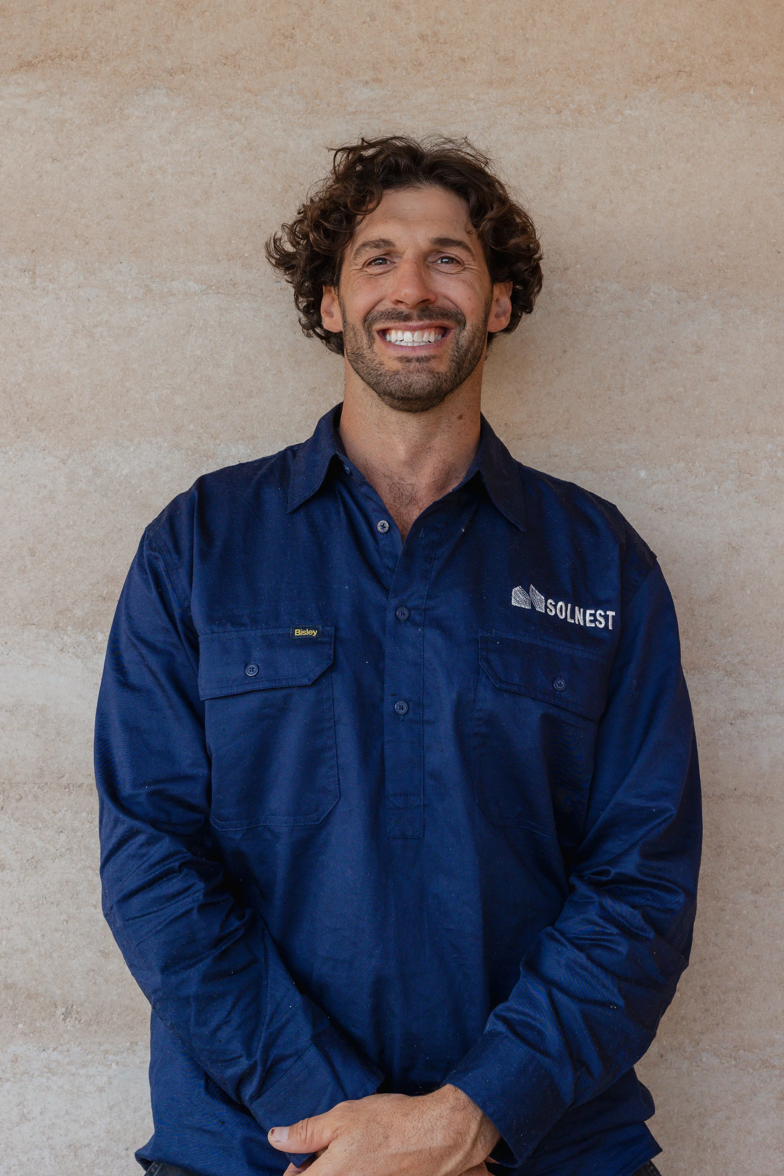 Man with dark curly hair and a beard smiling, wearing a navy blue shirt with SOLNEST logo.