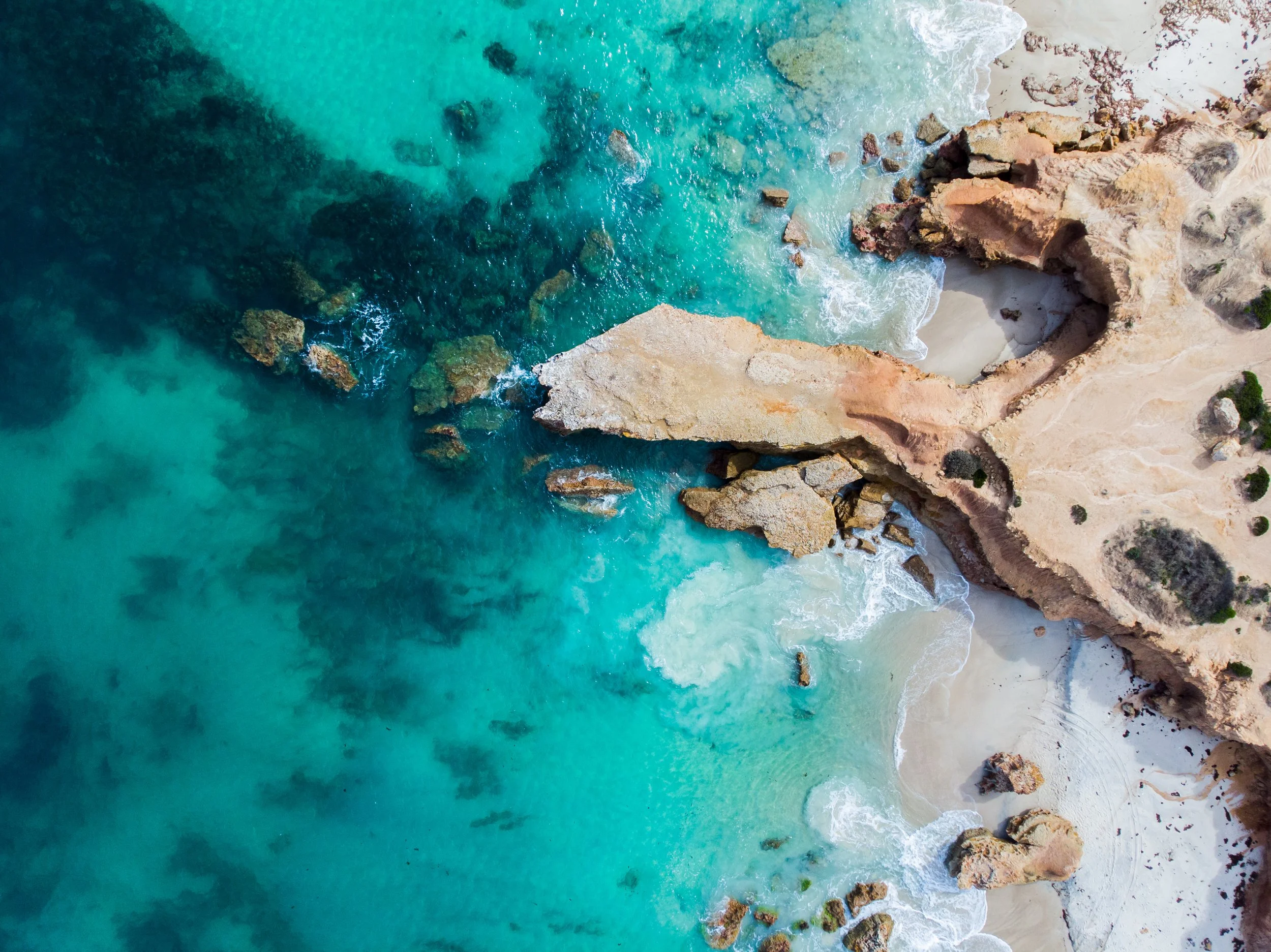 Aerial view of a rocky coastline with turquoise water crashing against the sandy shore and rocks.