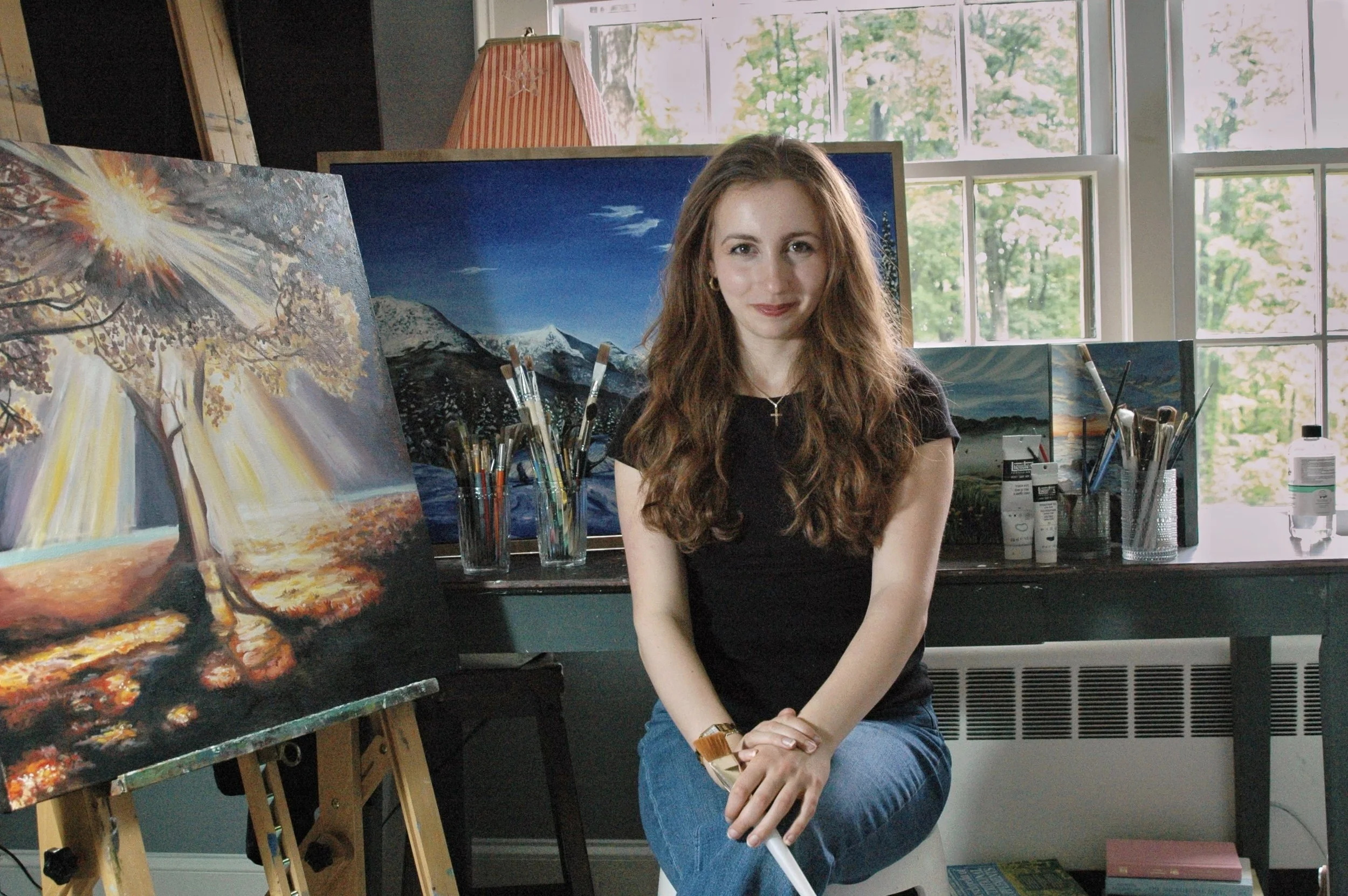 A young woman with long wavy brown hair sits in front of her landscape paintings in an art studio, holding a paintbrush and smiling at the camera.