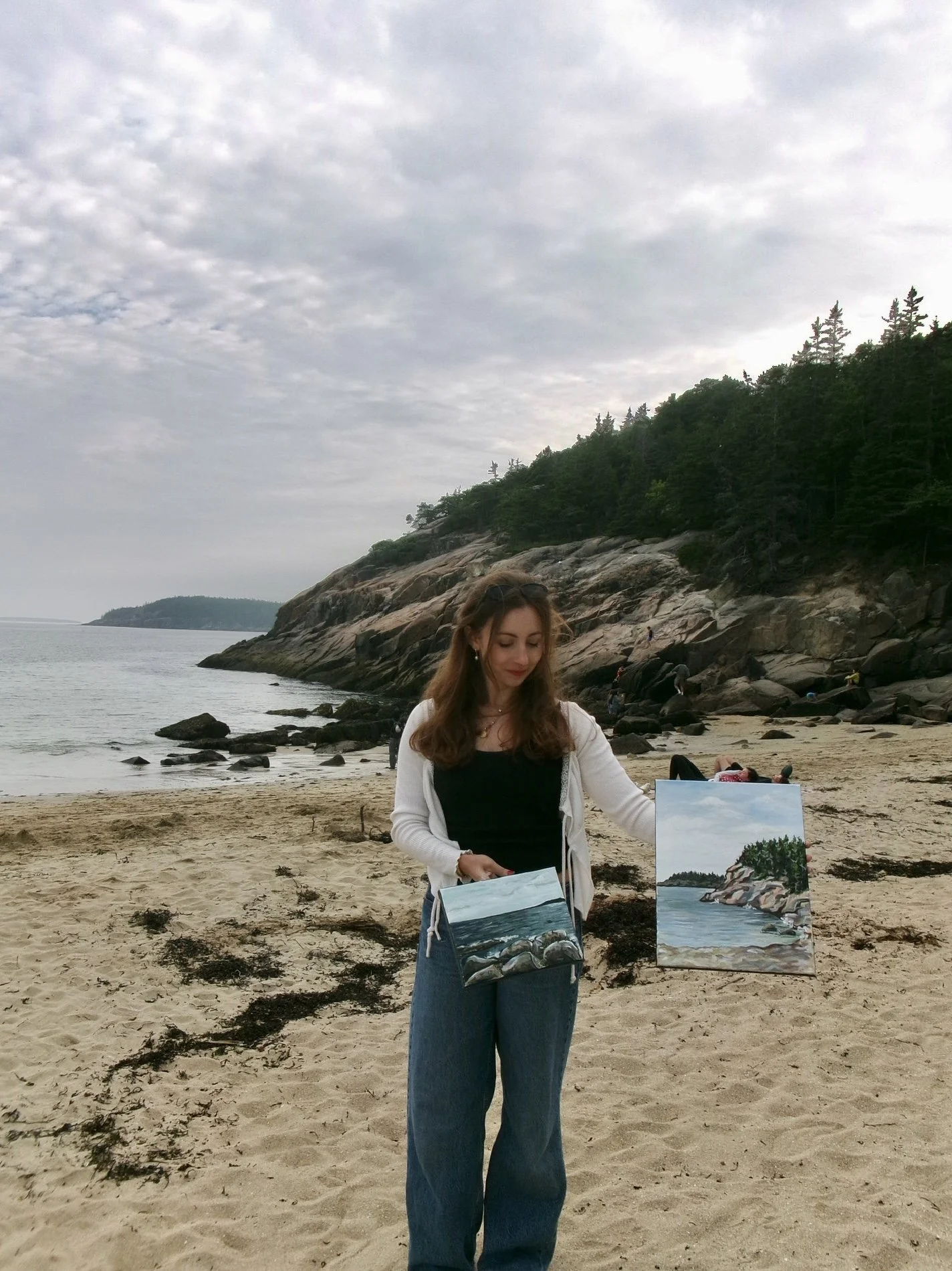 A woman on a beach holding two paintings of coastal landscapes, with rocky cliffs and the ocean in the background, and trees on the hillside.