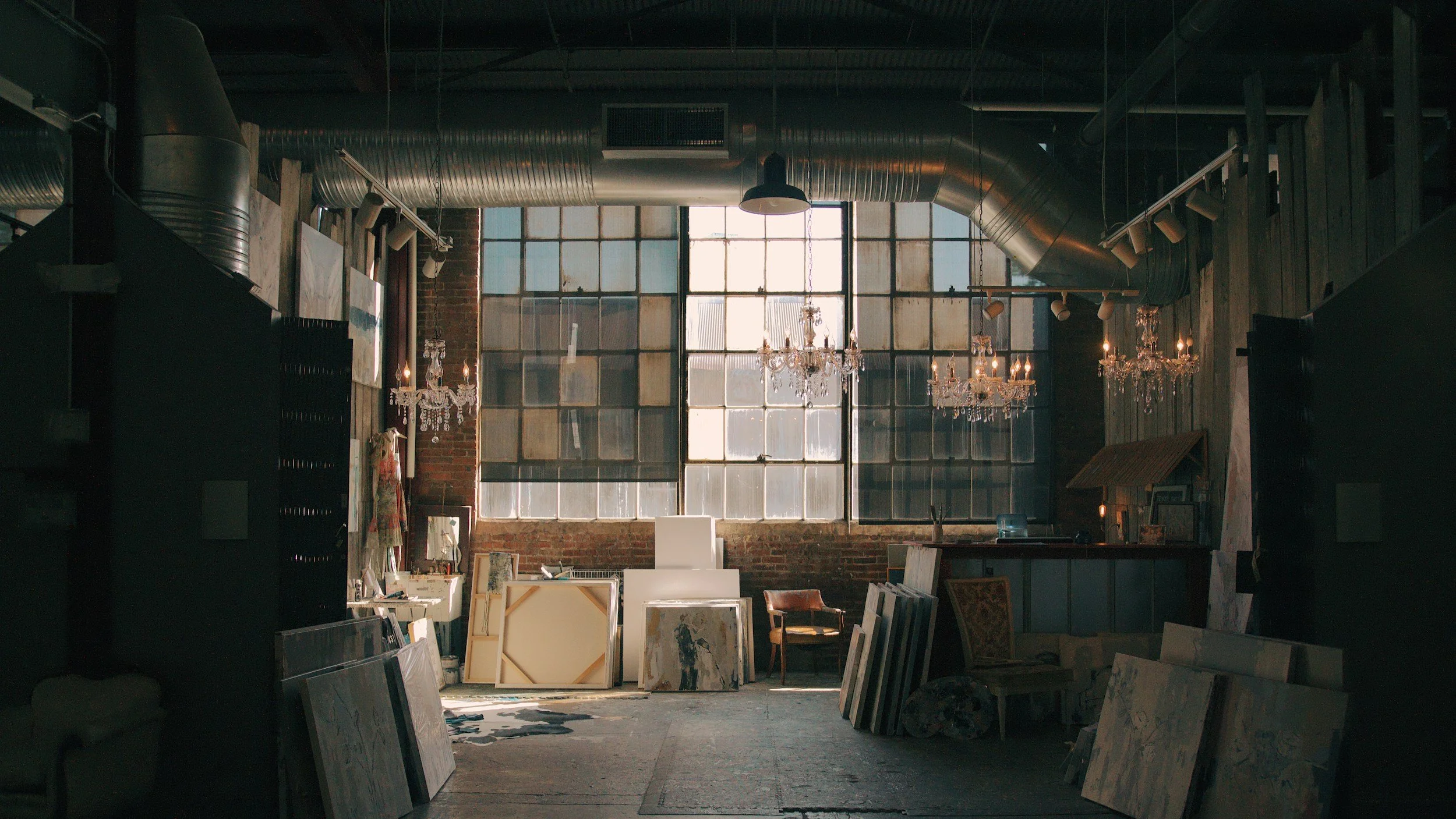 Interior of an art studio with large industrial windows, hanging chandeliers, and various paintings and canvases stored throughout.
